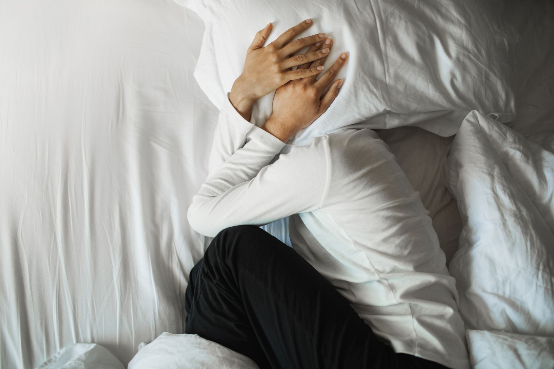 Frau liegt im Bett und bedeckt ihr Gesicht mit einem Kissen, wirkt gestresst oder verzweifelt.