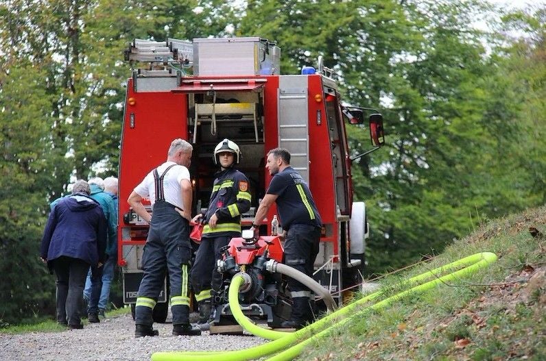 Drei Feuerwehrleute stehen an einem Feuerwehrauto mit Ausrüstung und gelben Schläuchen.