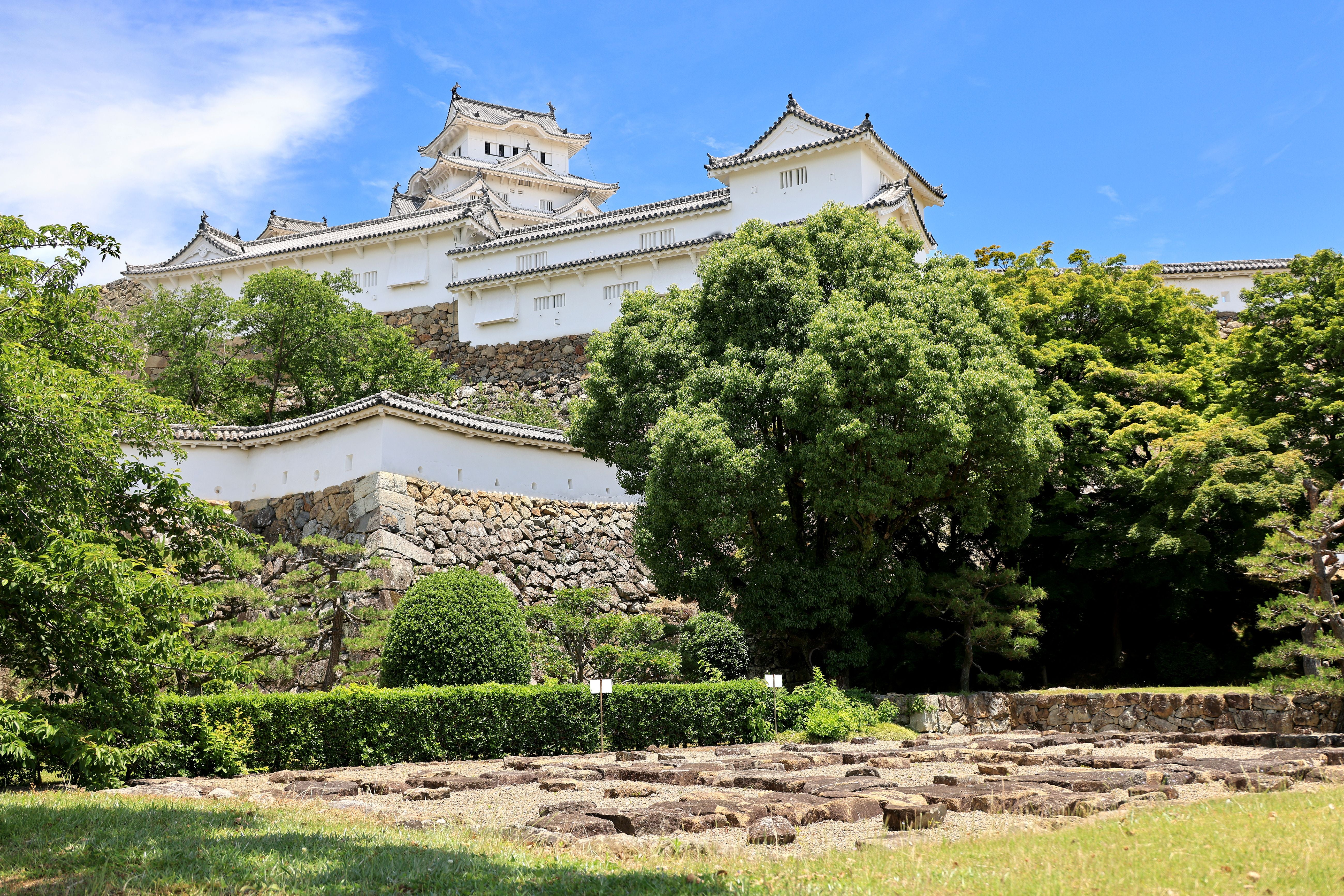 Himeji Schloss mit weißen Mauern, umgeben von Bäumen und Gartenruinen unter blauem Himmel.