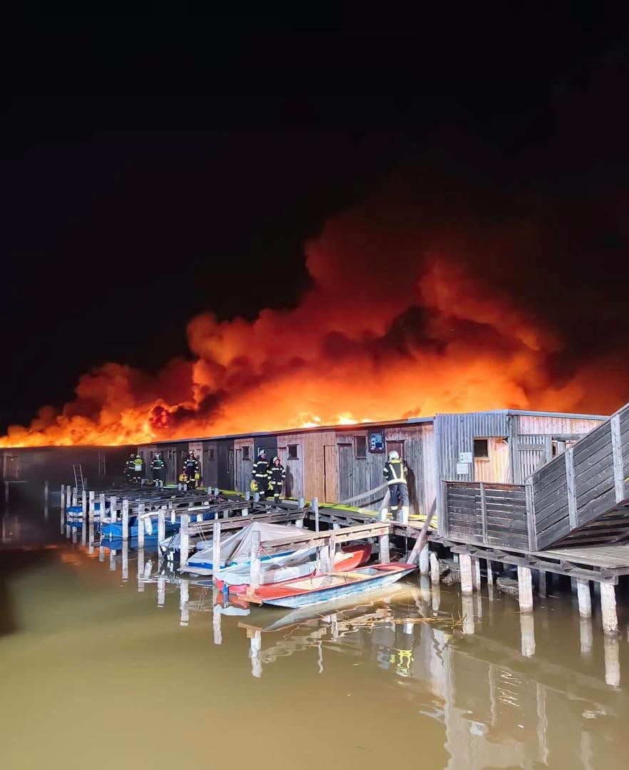 Großes Feuer bei Holzhütten am Wasser, Feuerwehrleute im Einsatz, starker Rauch am Nachthimmel.