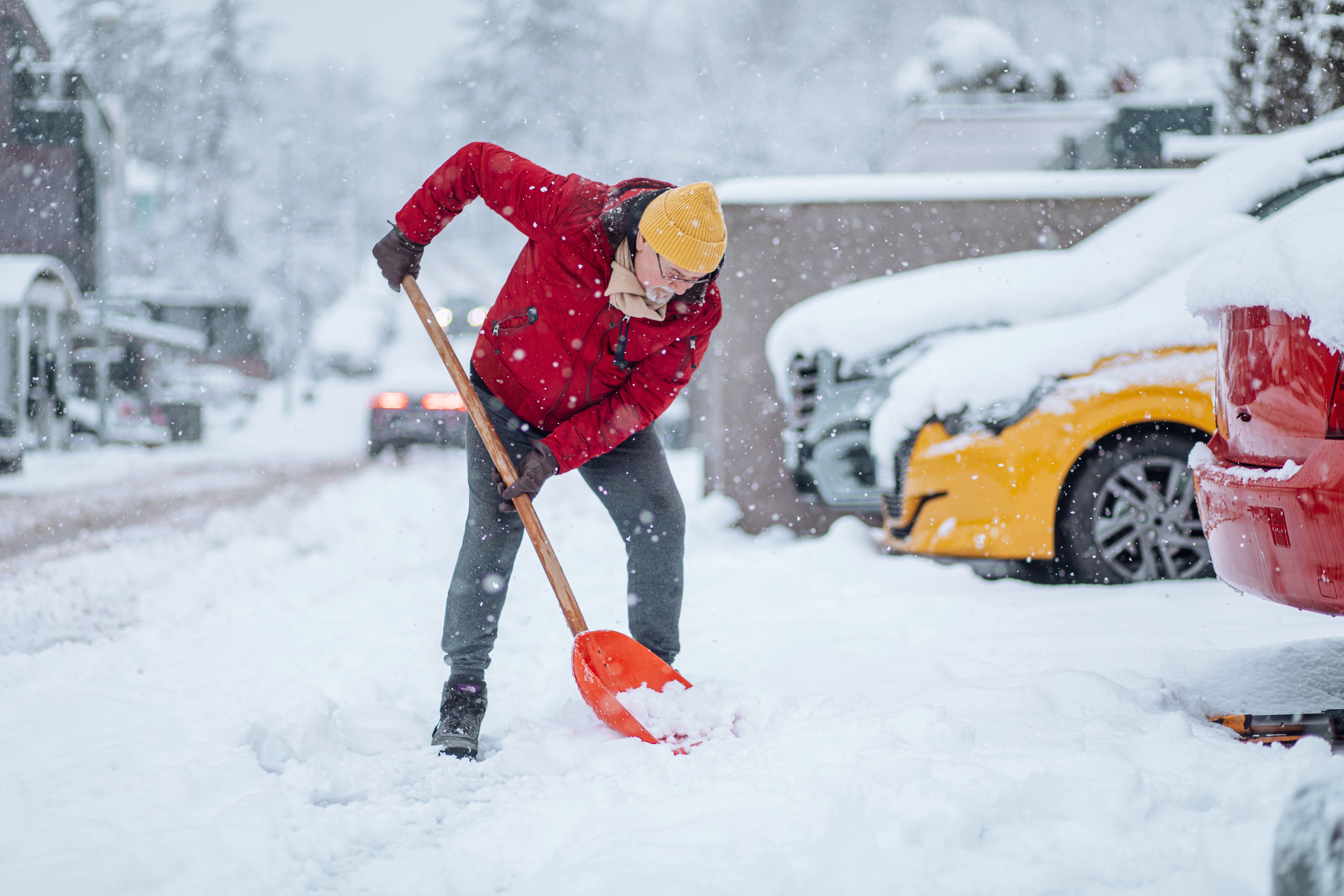 Person mit gelber Mütze und roter Jacke schaufelt Schnee von einer Einfahrt neben geparkten Autos.