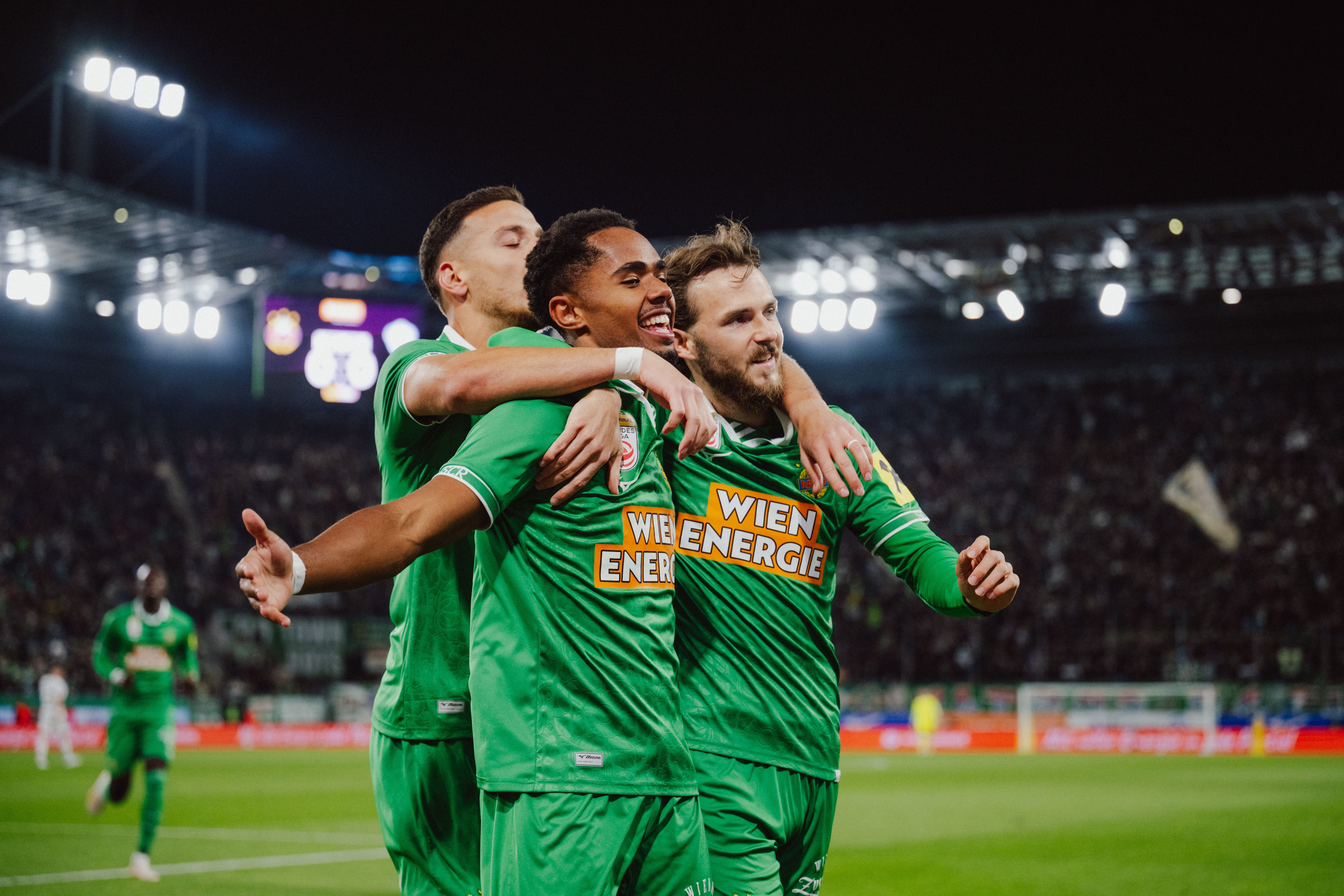 Petter Dahl jubelt strahlend im grünen Trikot und wird von seinen Rapid-Teamkollegen herzhaft umarmt.