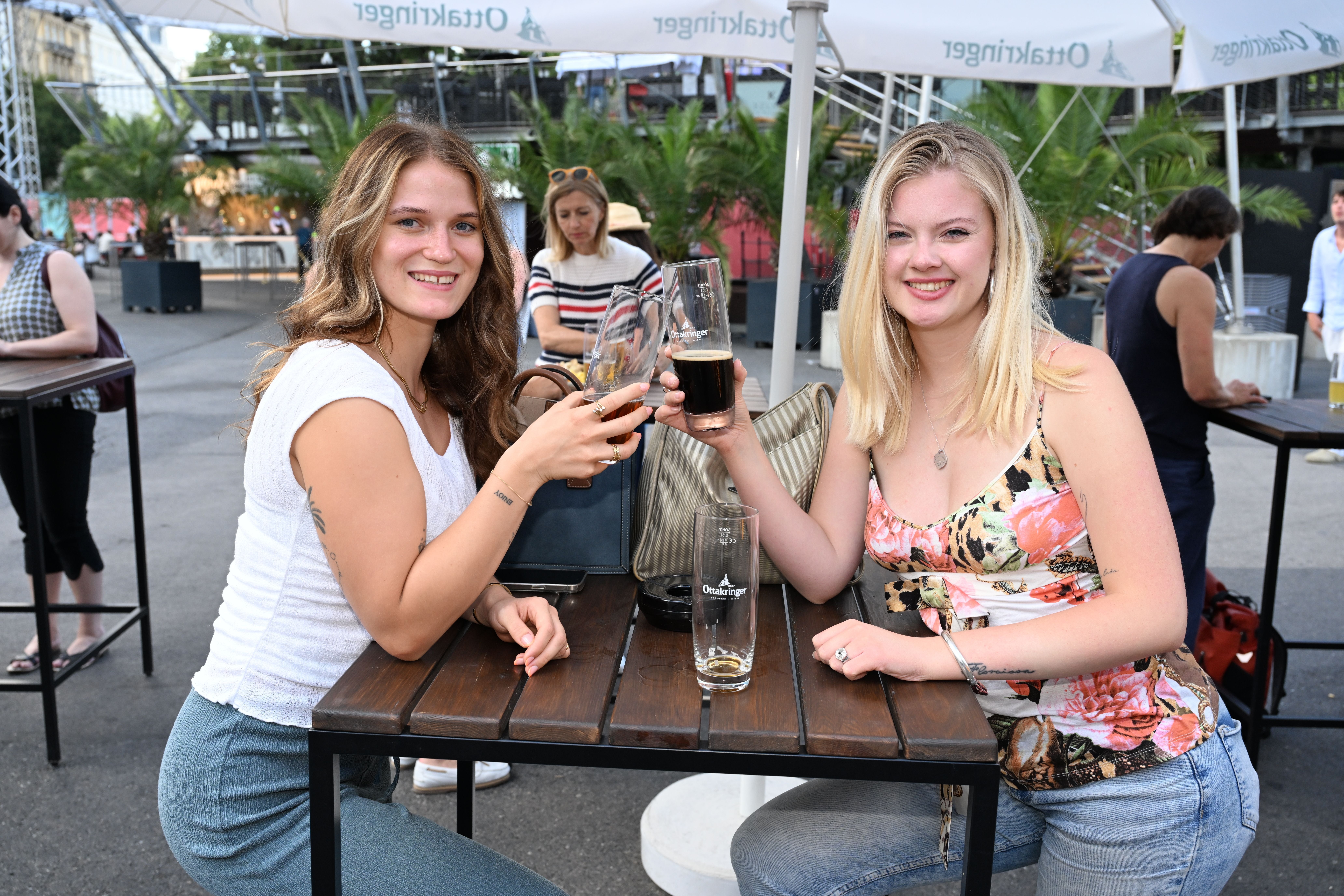 Zwei junge Frauen stoßen mit Biergläsern an einem Tisch im Freien beim Filmfestival an.