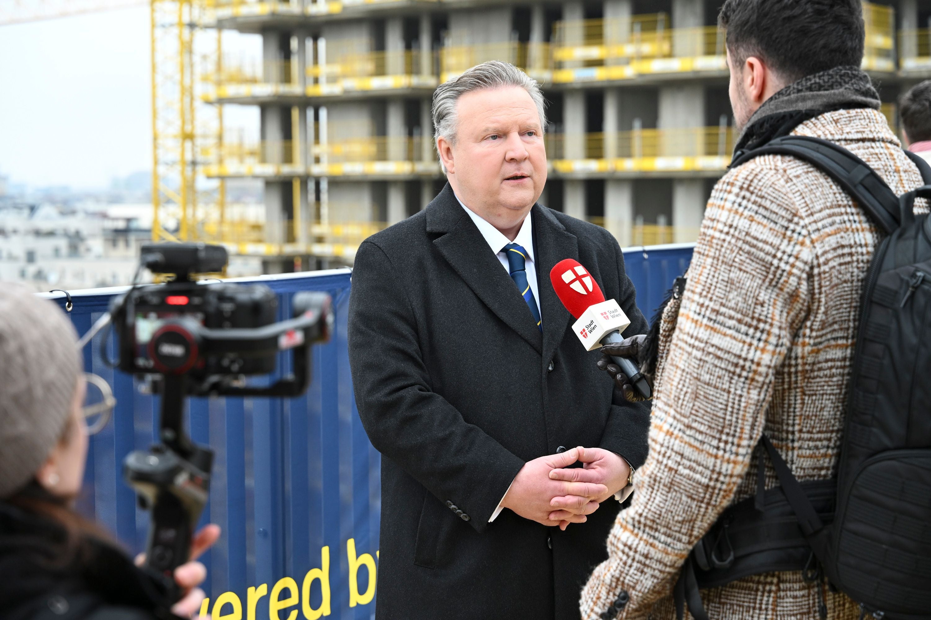 Ein Mann im Anzug gibt vor einer Baustelle ein Interview mit Mikrofon und Kamera.