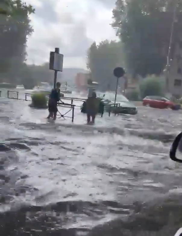 Straße in Rom überflutet, zwei Personen gehen durch das Wasser, Autos stehen im Hintergrund.