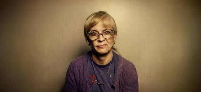 Frau mit Brille und lila Strickjacke sitzt vor neutralem Hintergrund und blickt in die Kamera.