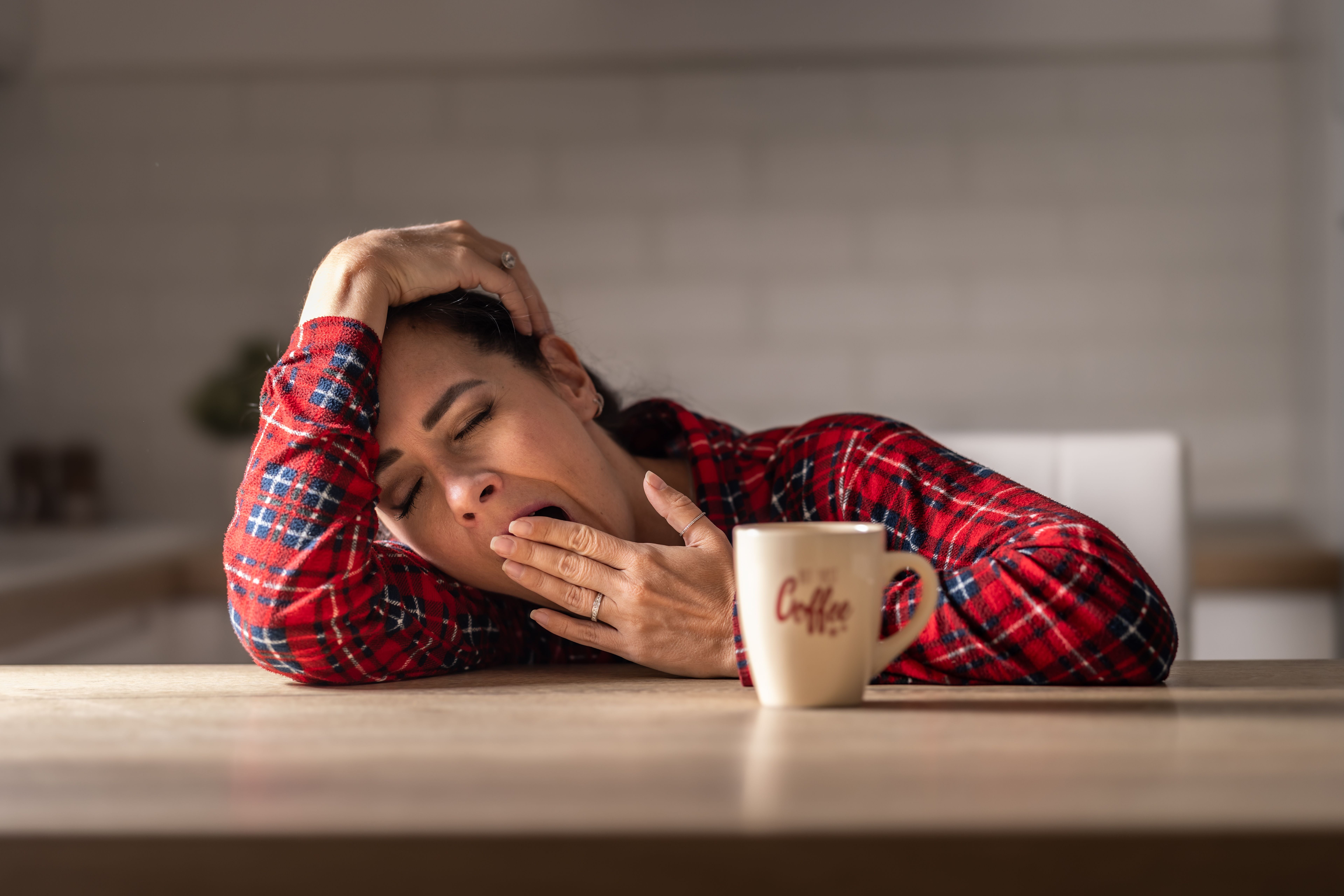 Frau im roten Hemd gähnt müde am Tisch, vor ihr steht eine Tasse mit der Aufschrift „Coffee“.
