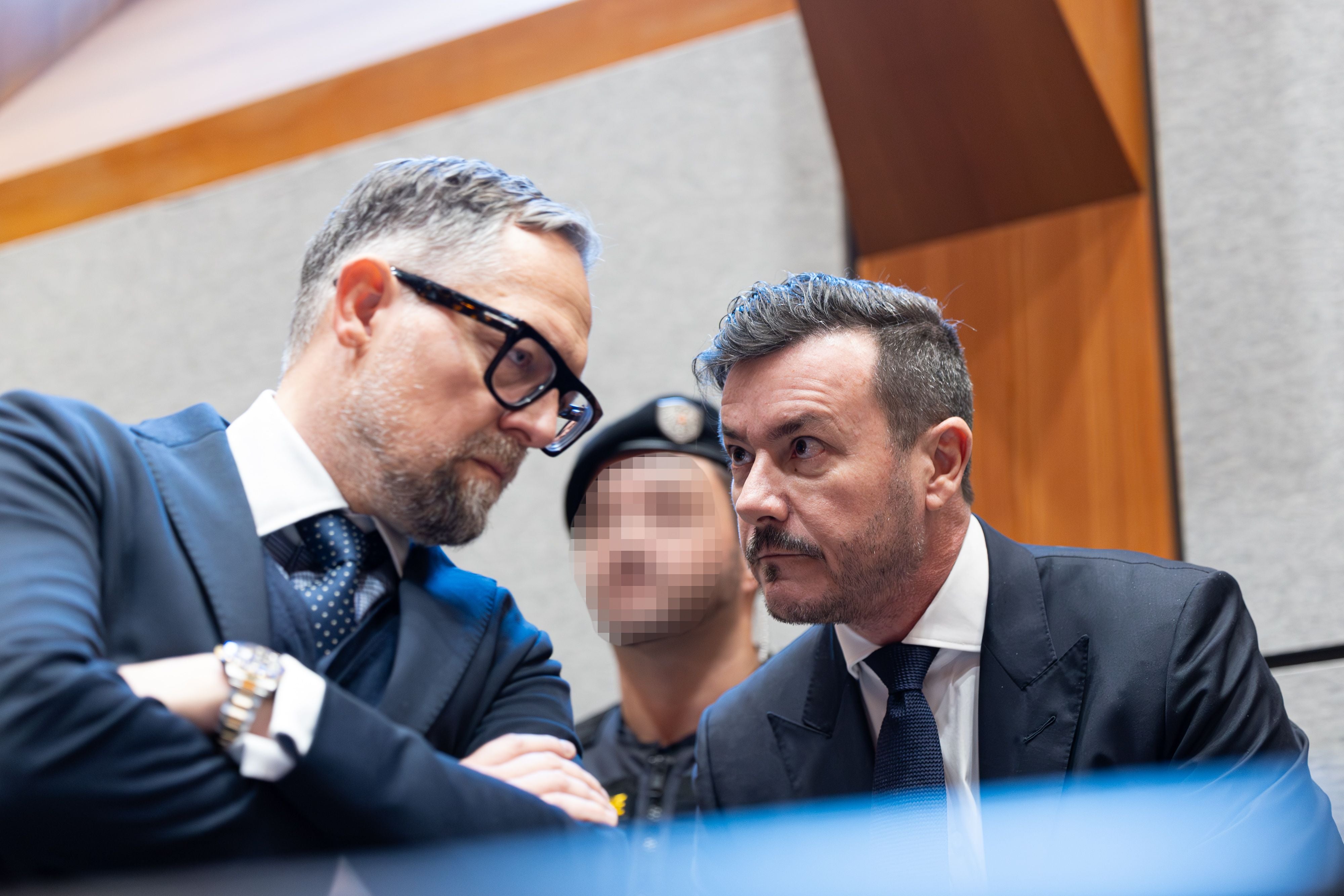 Zwei Männer im Anzug sprechen sich ernsthaft in einem Gerichtssaal ab, eine Person im Hintergrund unscharf.