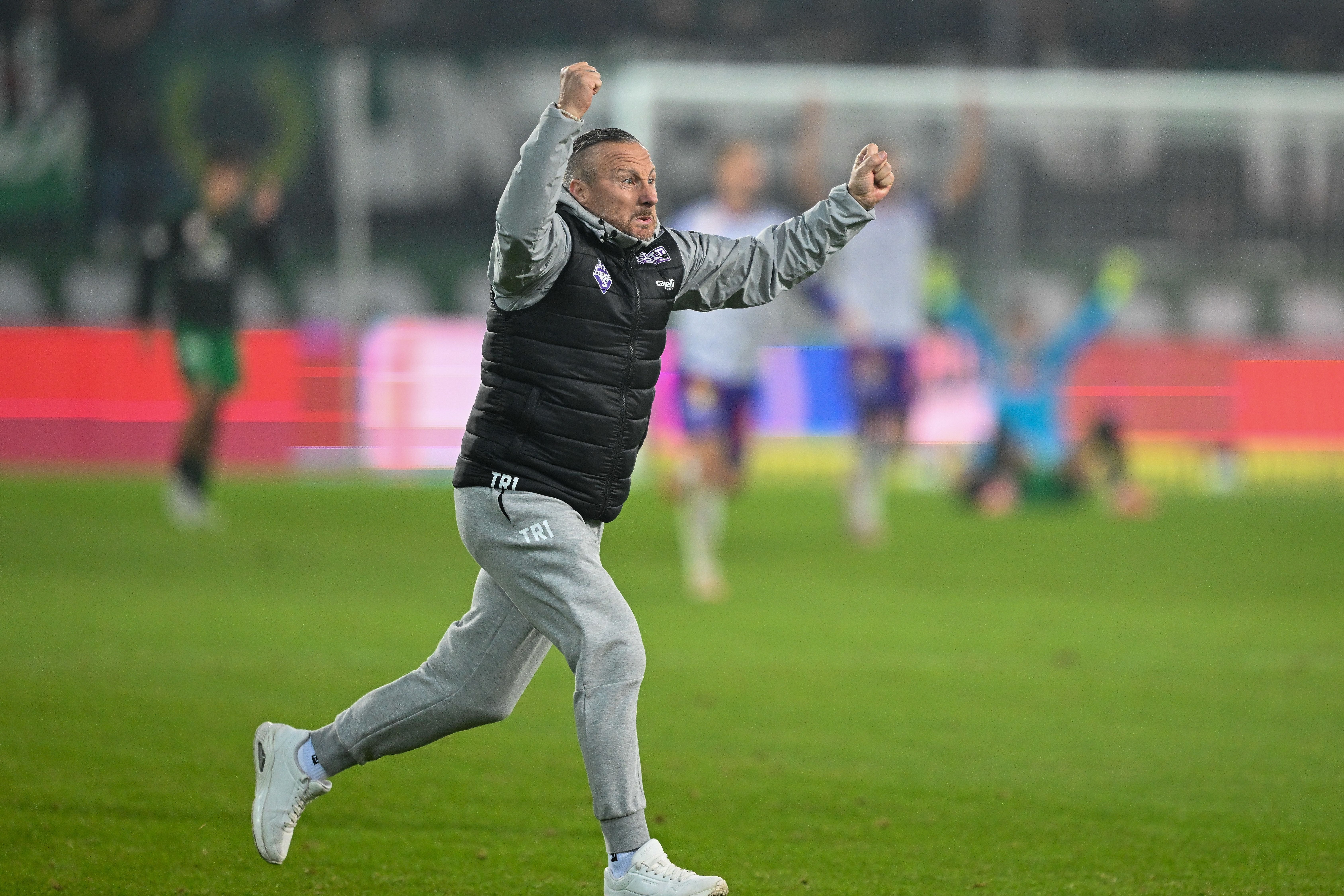 Ein Trainer im Trainingsanzug jubelt mit erhobenen Fäusten auf dem Fußballfeld.
