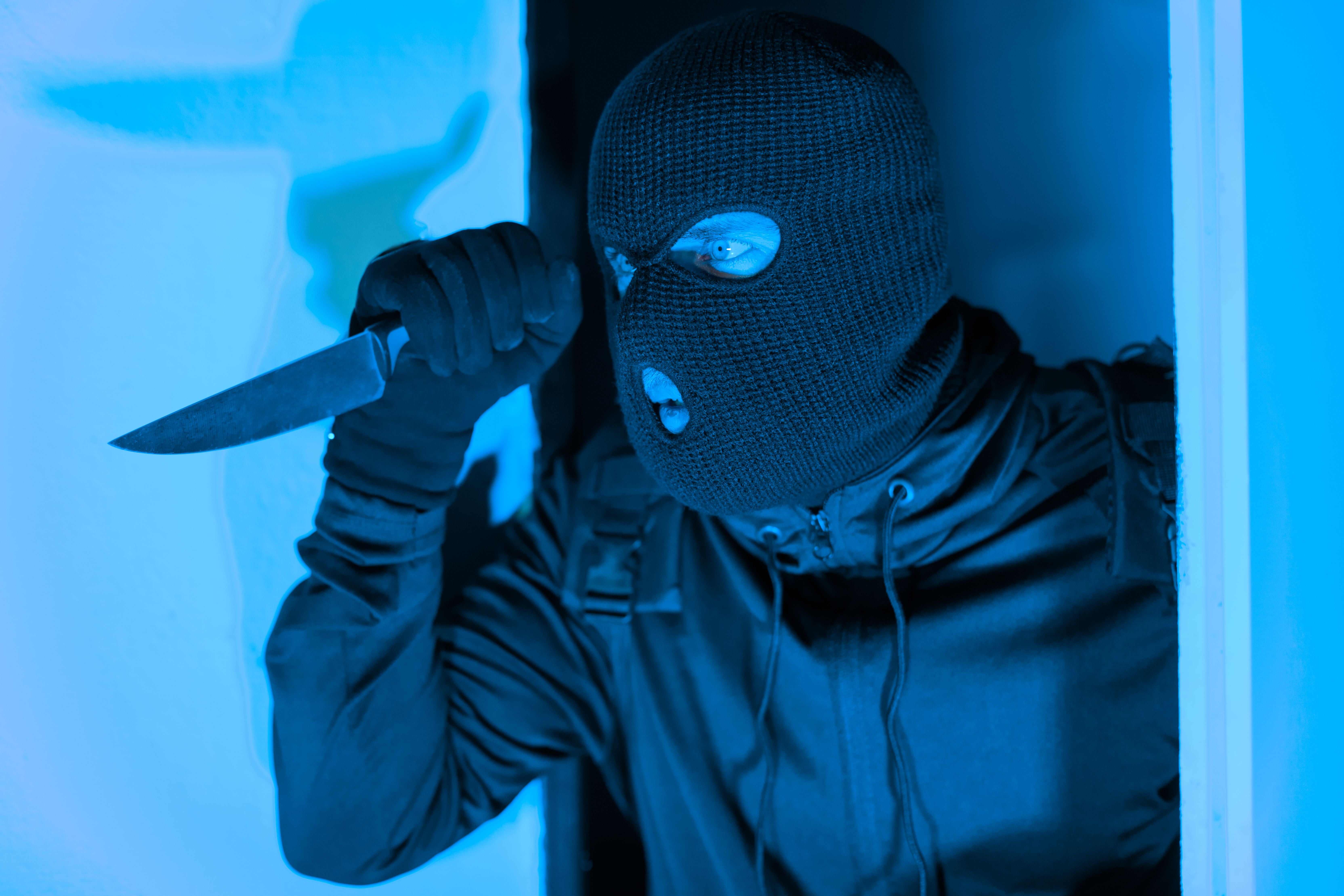 Maskierter Einbrecher mit erhobenem Messer in einem Türrahmen, bedroht eine Wohnung.