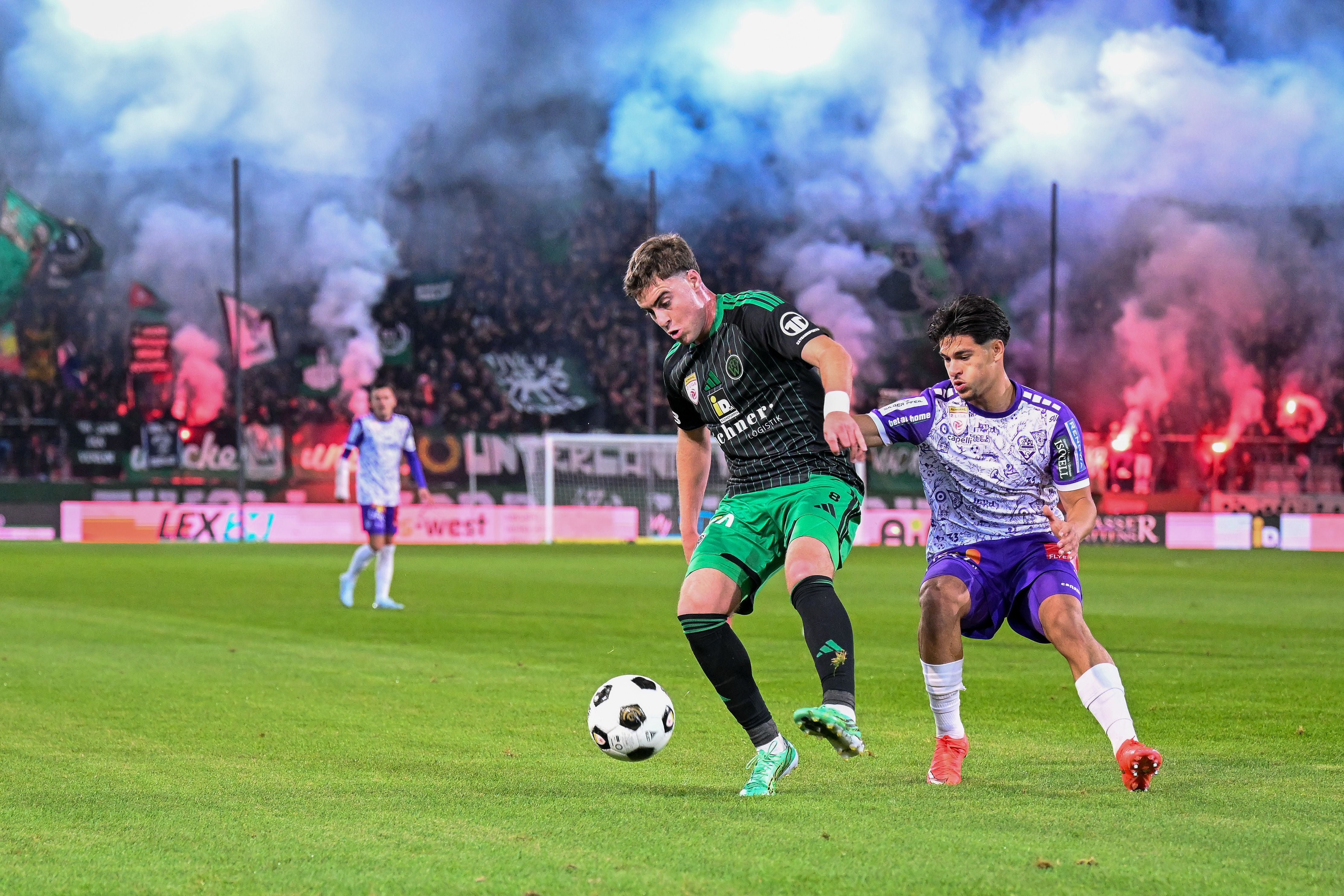 Zwei Fußballspieler kämpfen im Stadion um den Ball, im Hintergrund Rauch und jubelnde Fans.
