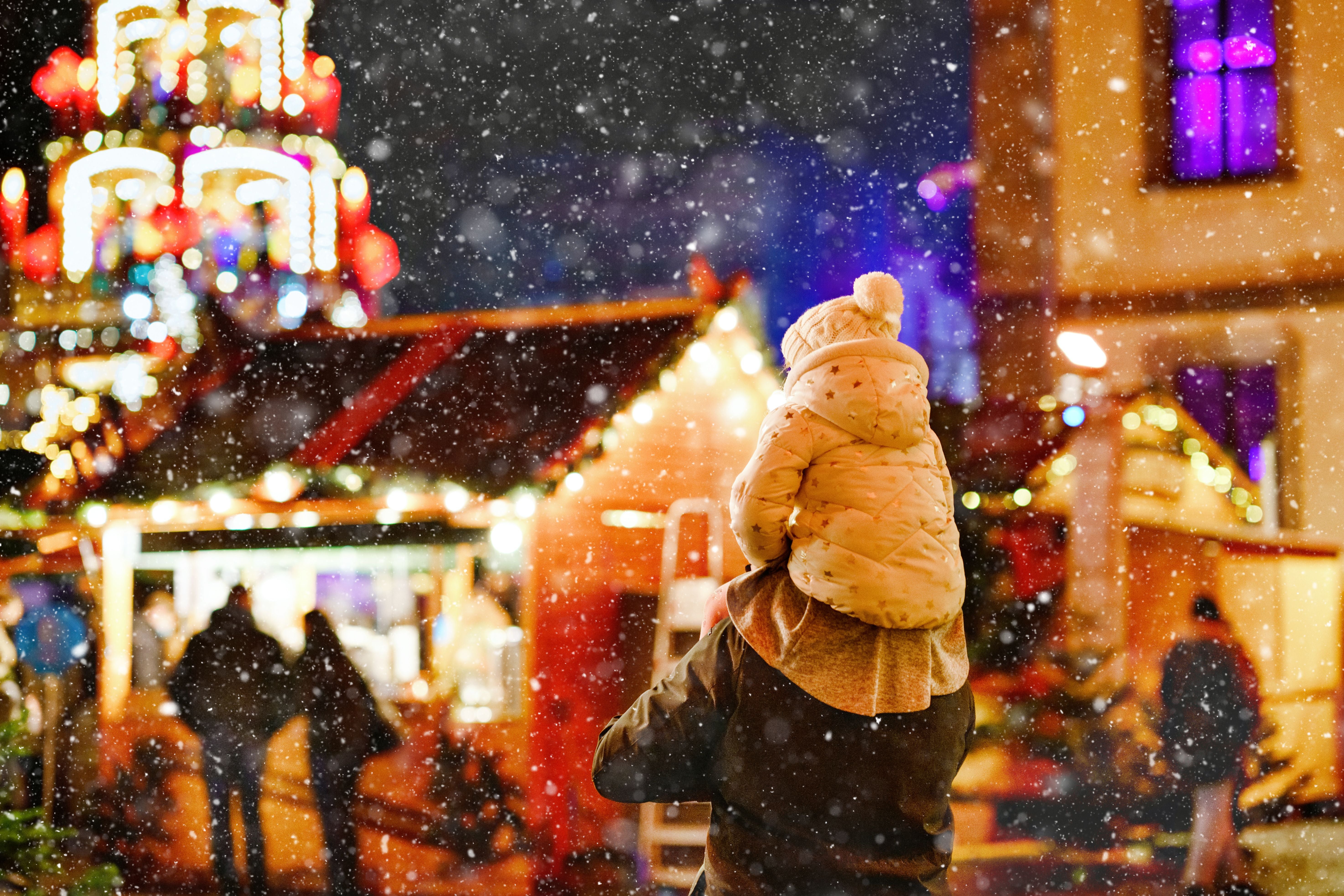 Vater trägt kleines Kind auf den Schultern auf einem verschneiten, beleuchteten Weihnachtsmarkt.