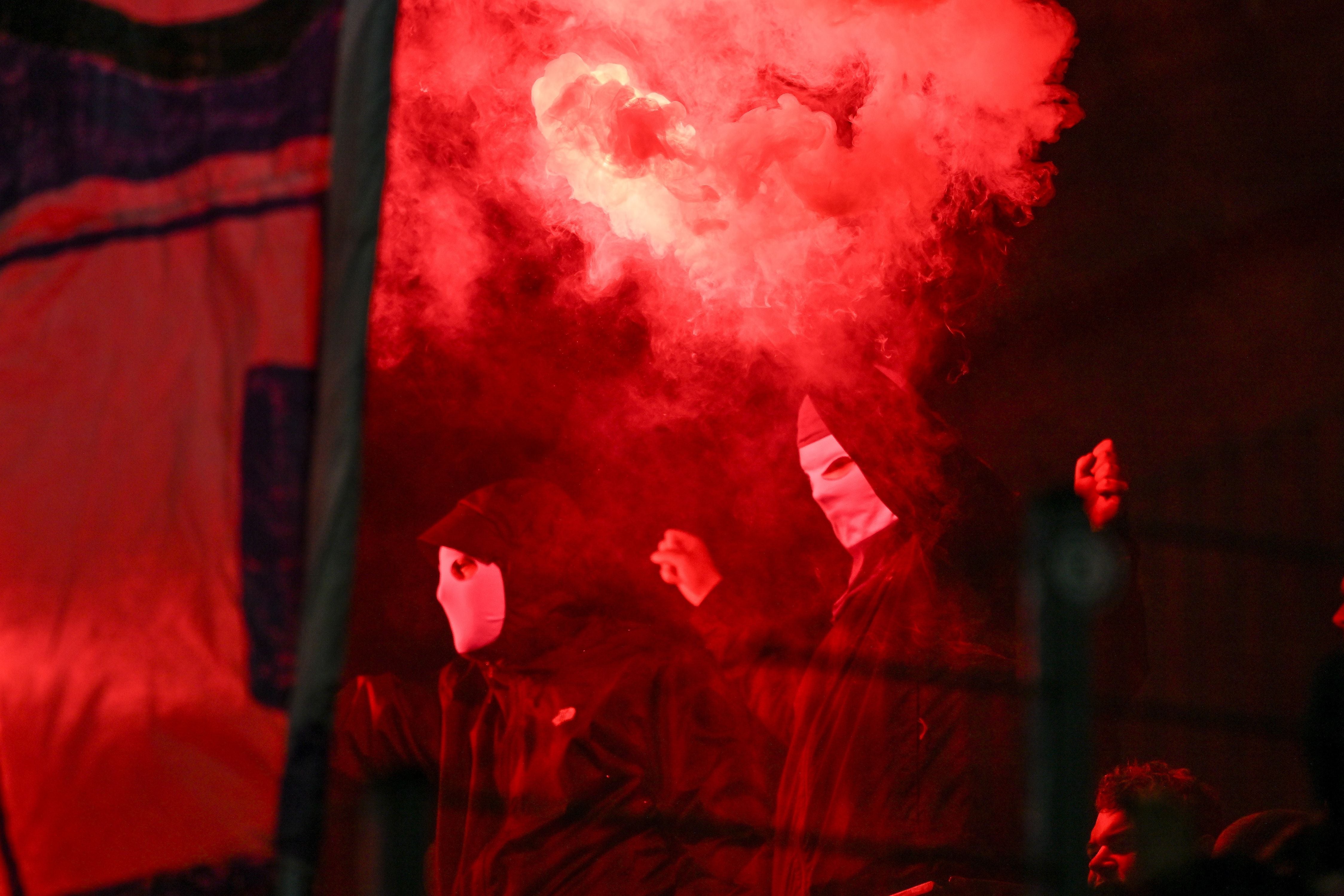 Zwei vermummte Fans mit Masken stehen im roten Rauch auf der Tribüne.