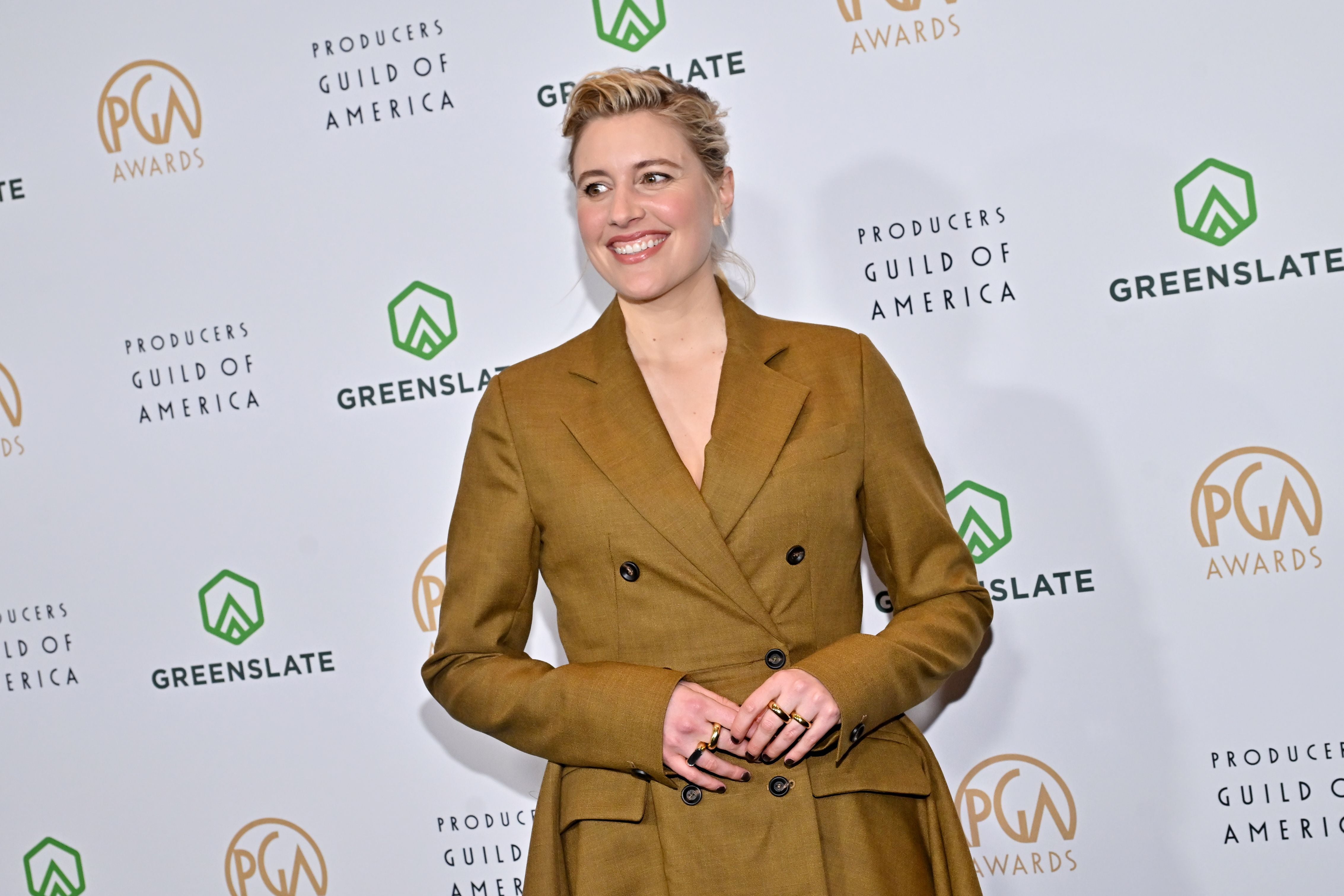 Frau in braunem Anzug lächelt auf rotem Teppich bei den PGA Awards vor Sponsorenwand.