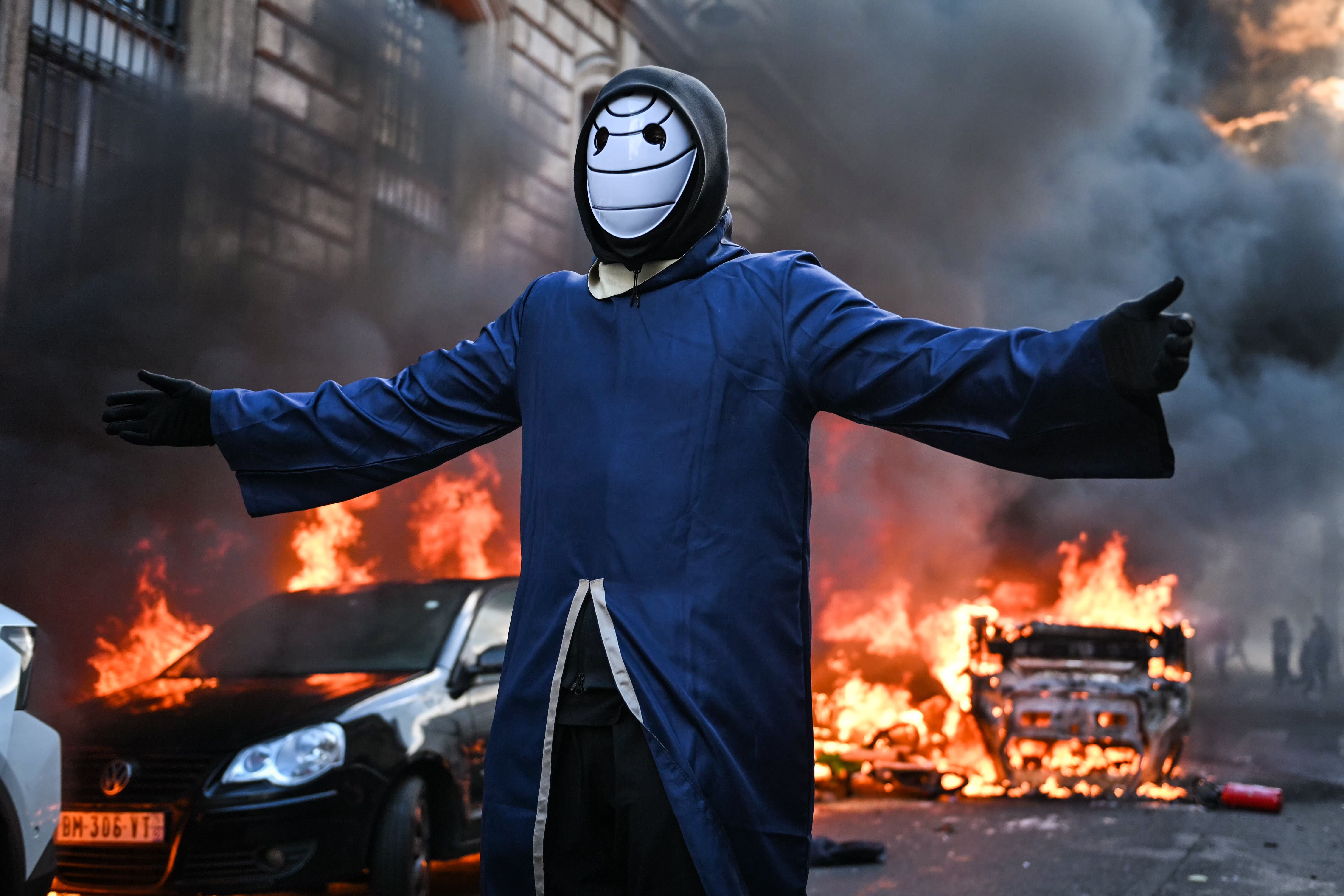 Person mit Maske steht mit ausgebreiteten Armen vor brennenden Autos und Rauch in Paris.