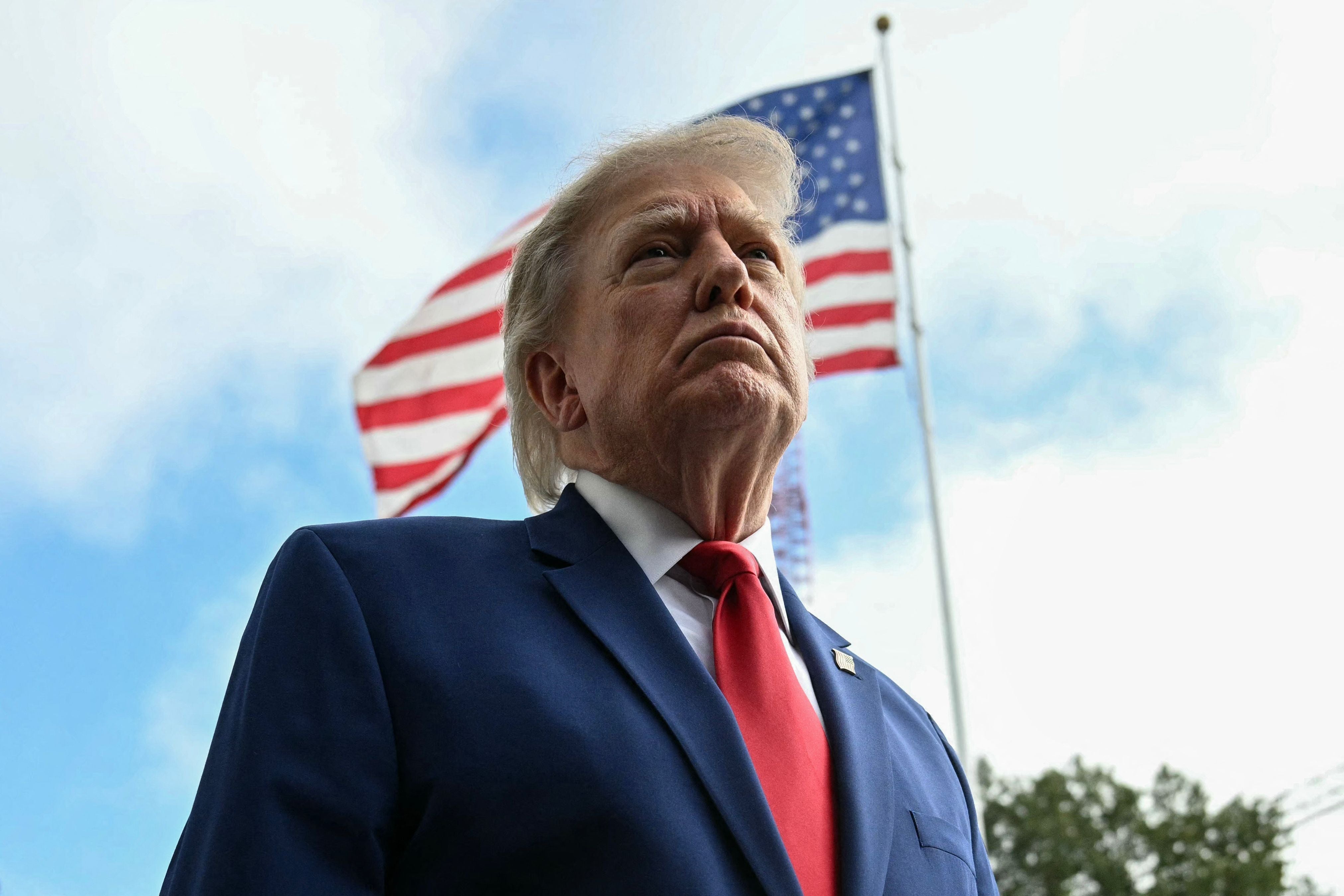 Mann im blauen Anzug mit roter Krawatte steht vor wehender US-Flagge und blickt ernst nach oben.