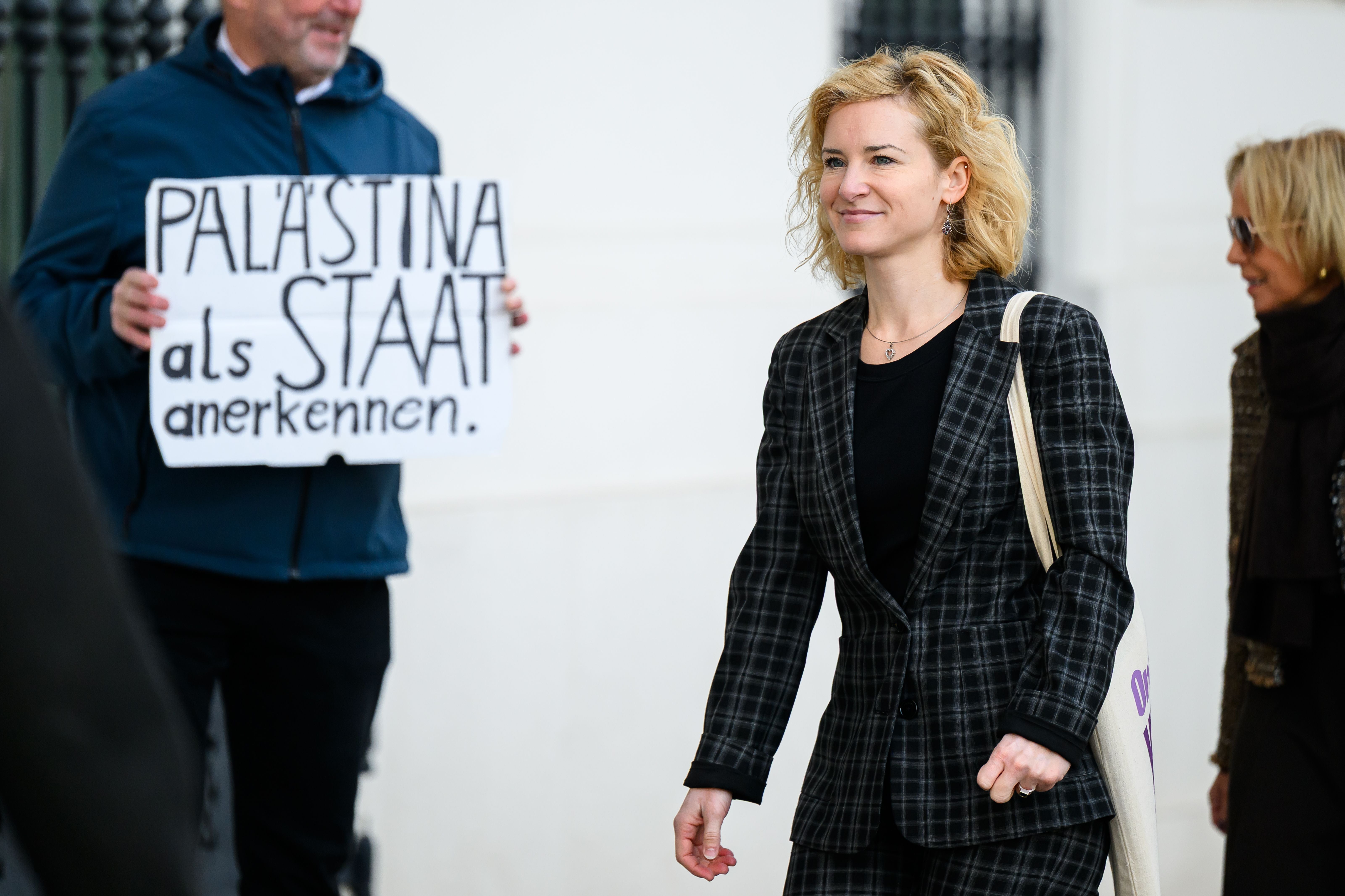 Eine Frau im karierten Anzug geht vorbei, im Hintergrund hält jemand ein Pro-Palästina-Plakat.