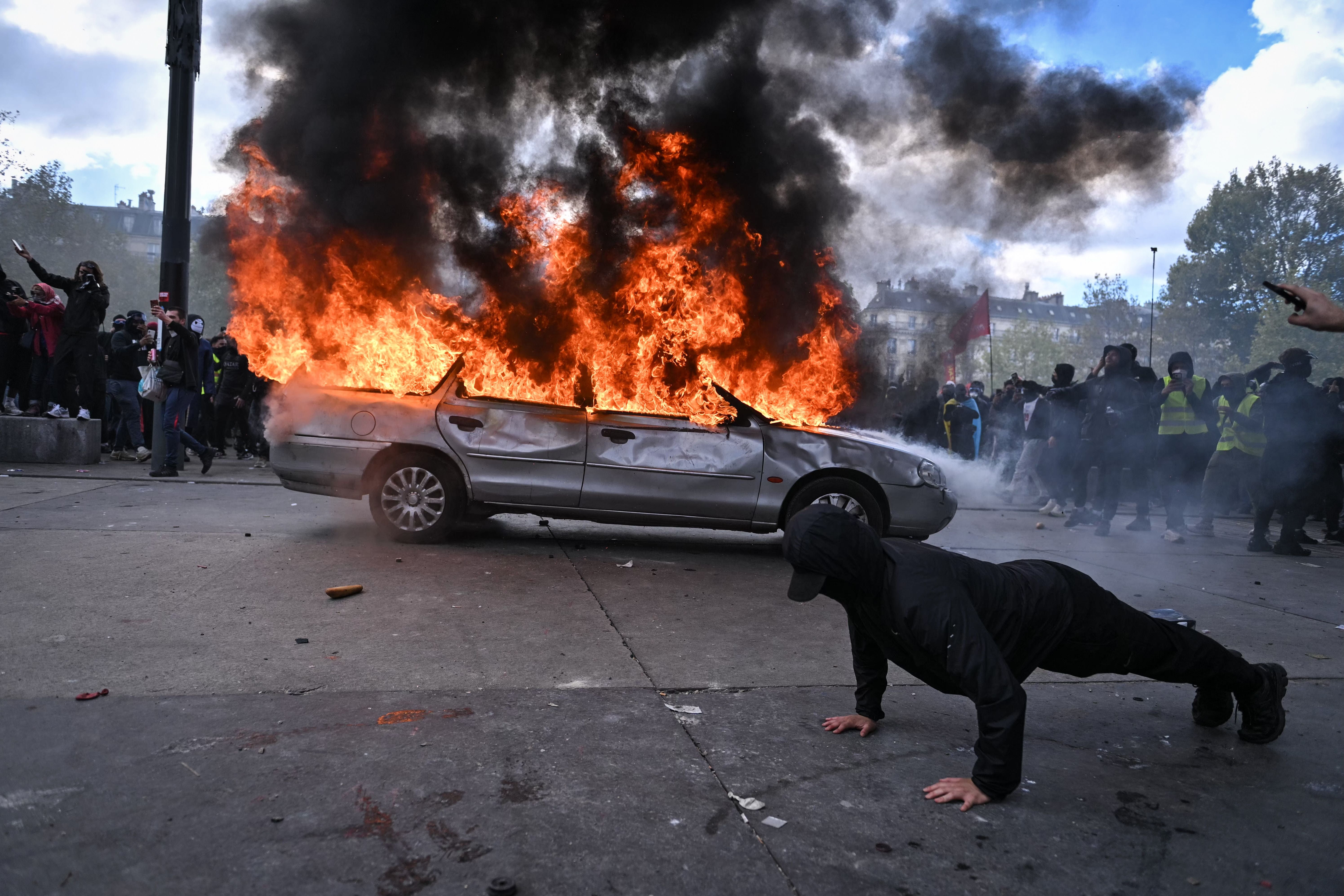 Ein Auto steht in Flammen, während Menschen und ein Mann beim Bodenstütz im Vordergrund protestieren.
