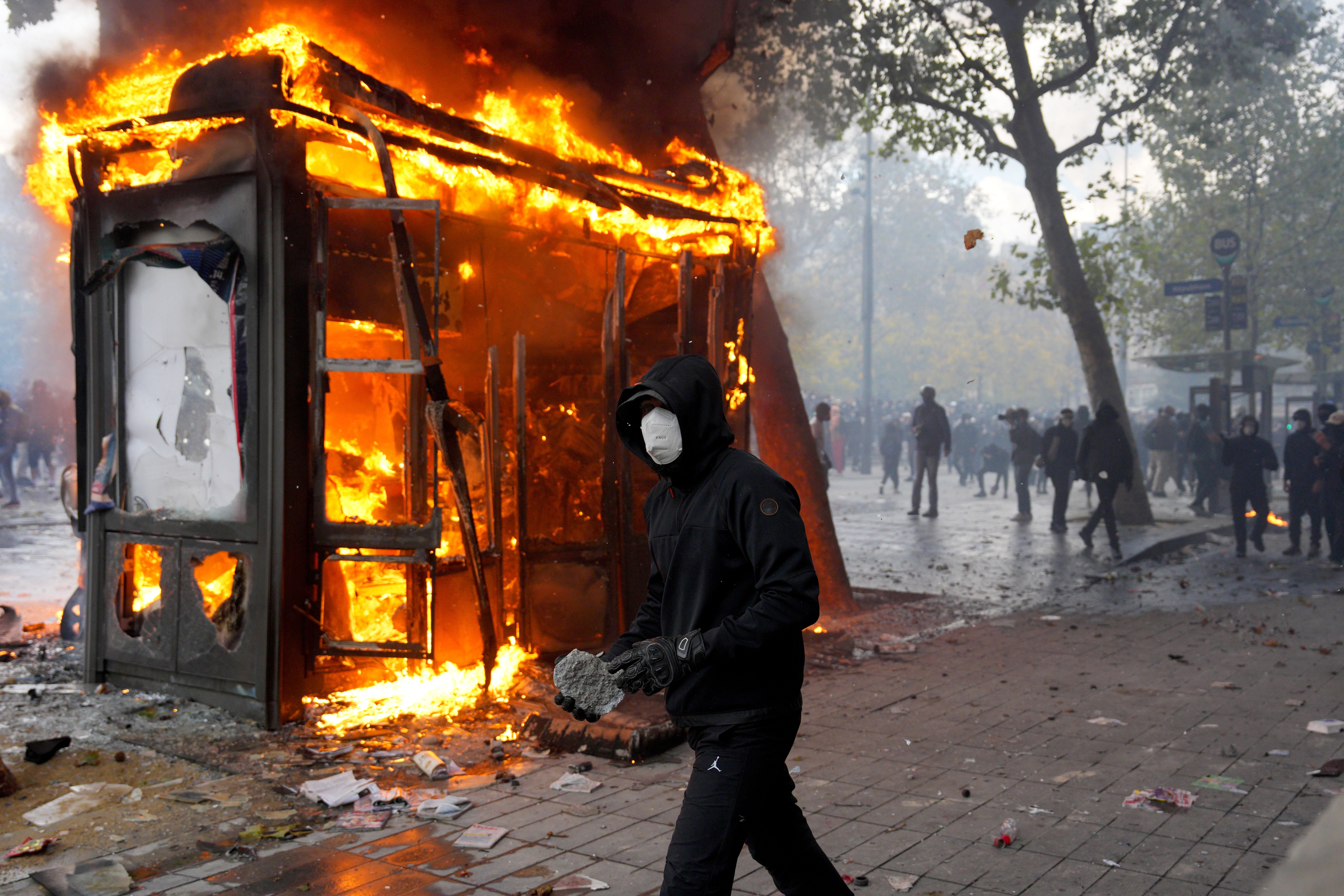Ein vermummter Demonstrant hält einen Stein, während im Hintergrund ein Kiosk in Flammen steht.