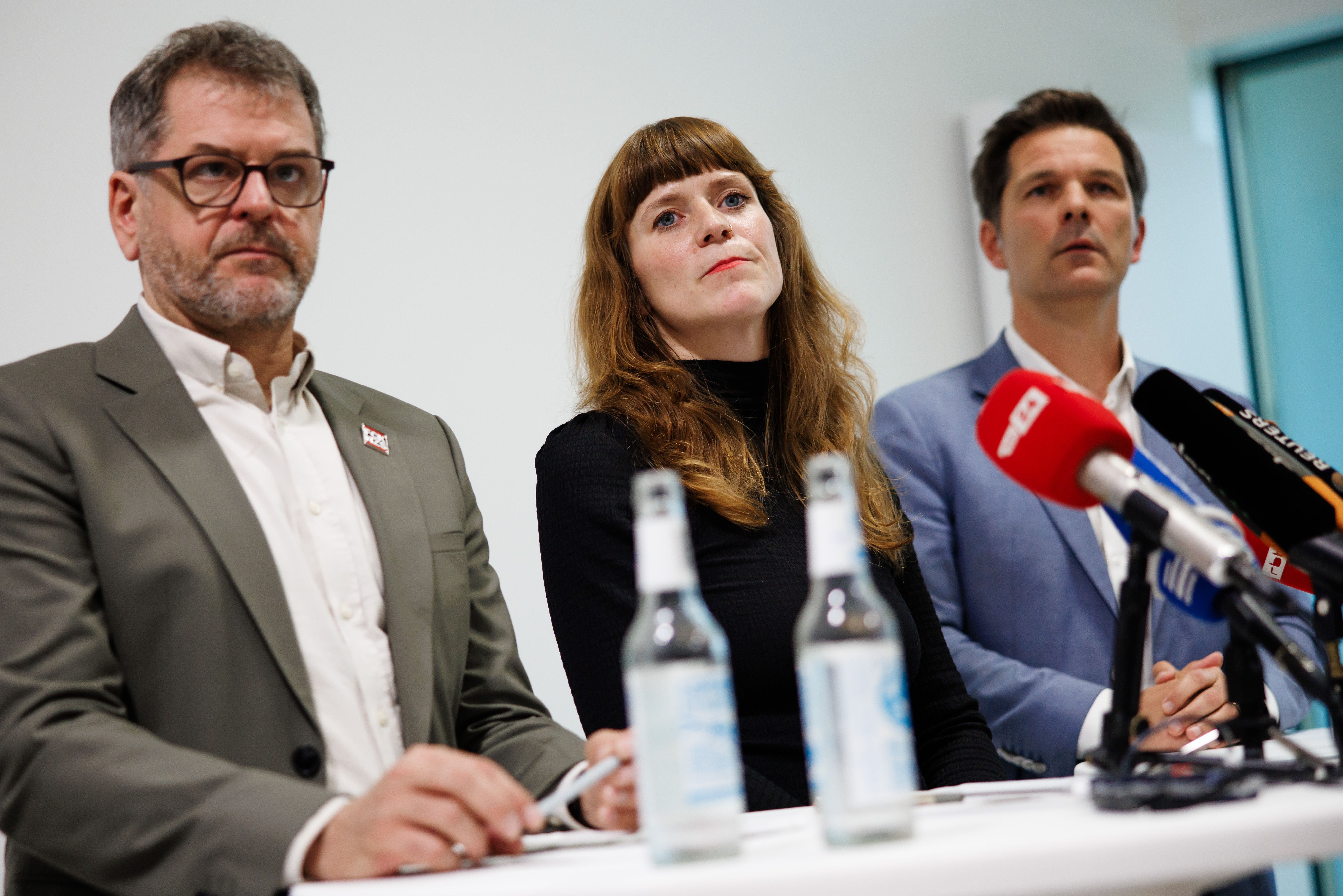 Drei Personen sitzen auf einer Pressekonferenz mit Mikrofonen und Wasserflaschen auf dem Tisch.