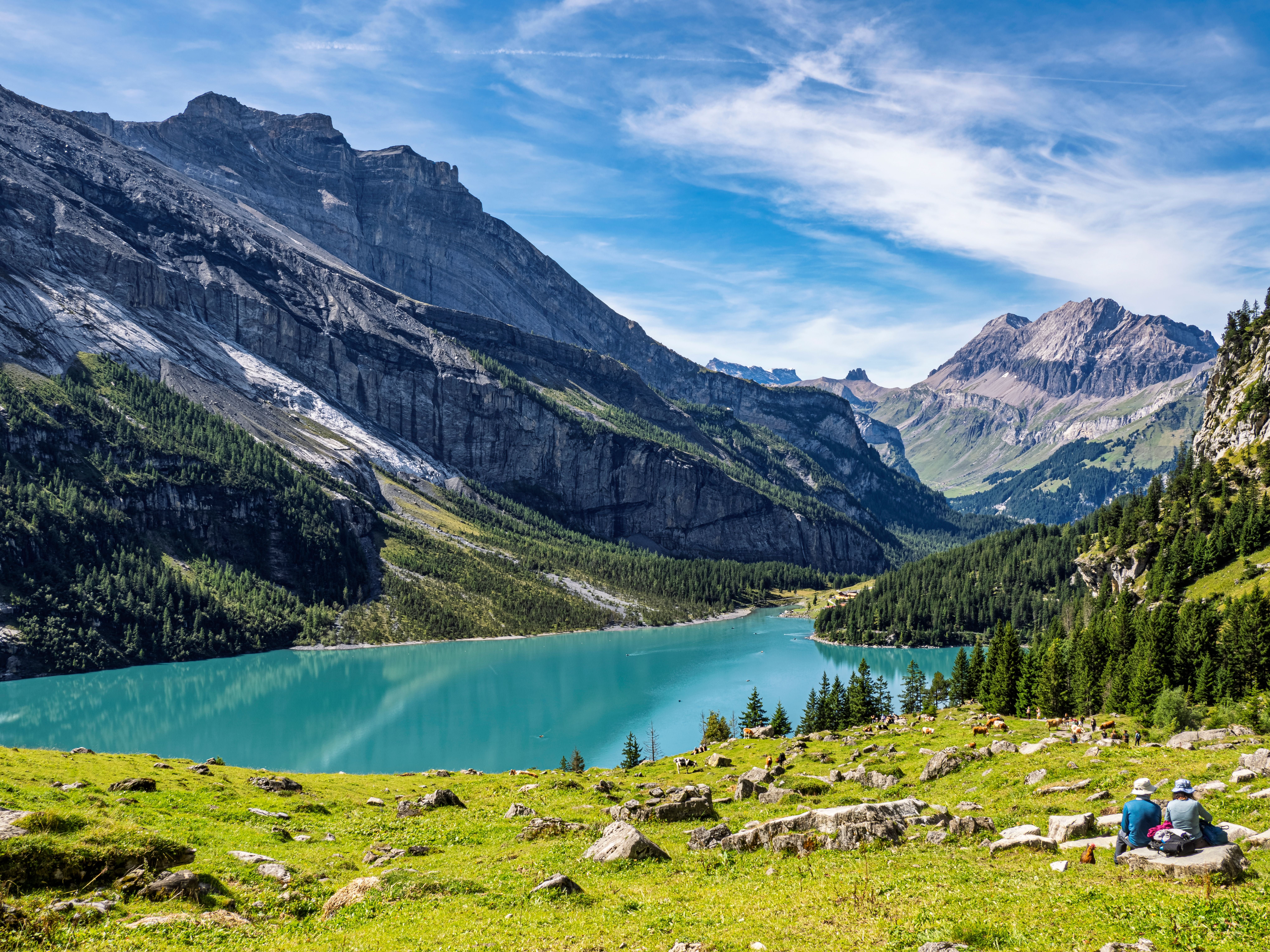 Blick auf den türkisblauen Oeschinensee vor majestätischen Alpen, zwei Personen sitzen im Vordergrund.