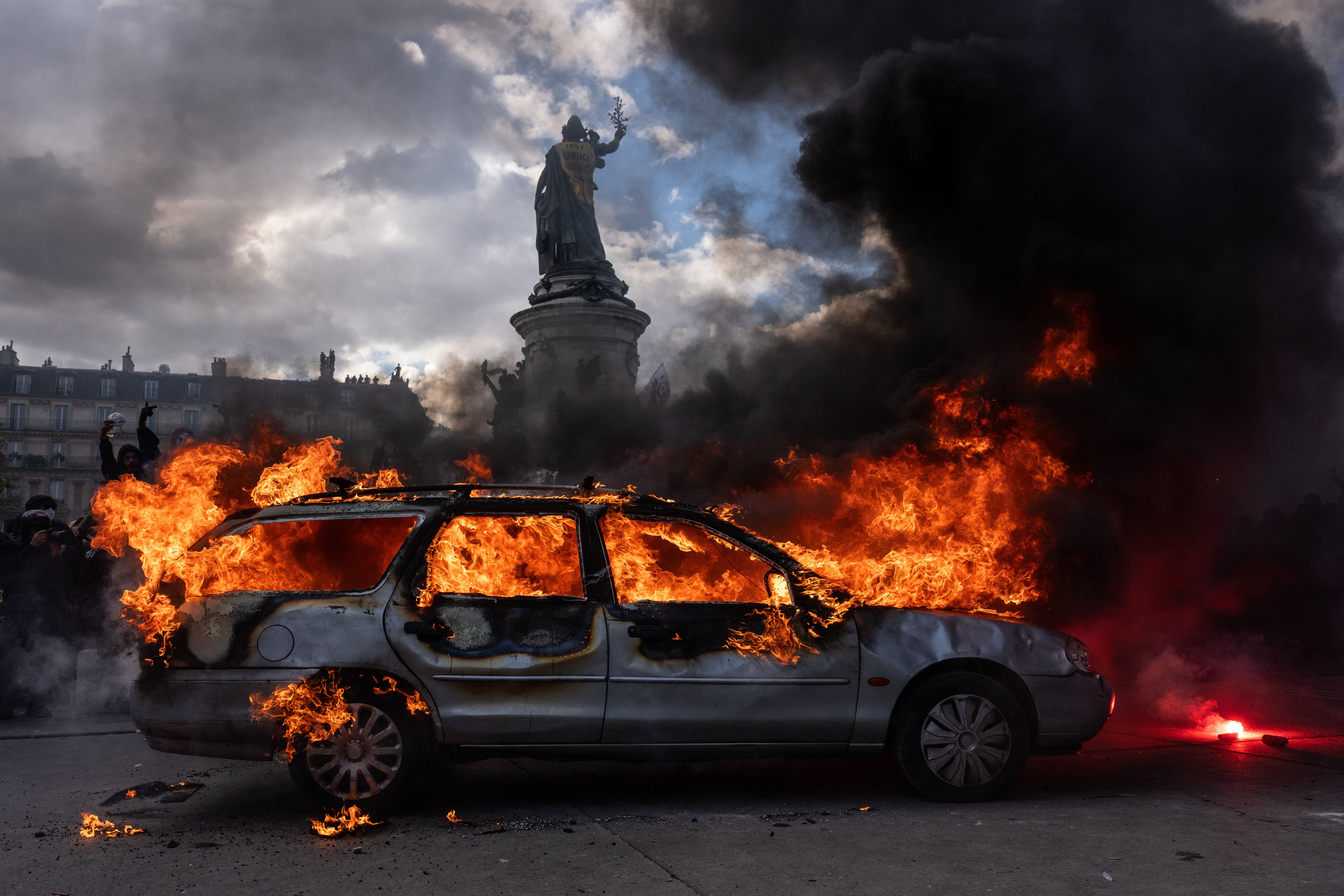 Ein Auto brennt lichterloh auf einem Platz, dichter schwarzer Rauch steigt auf, im Hintergrund eine Statue.