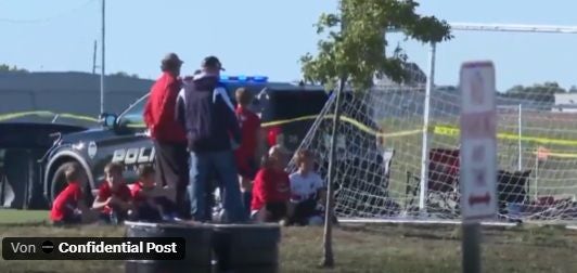 Kinder in roten Trikots sitzen neben Erwachsenen und einem Polizeiauto auf einem Sportfeld in Wichita.