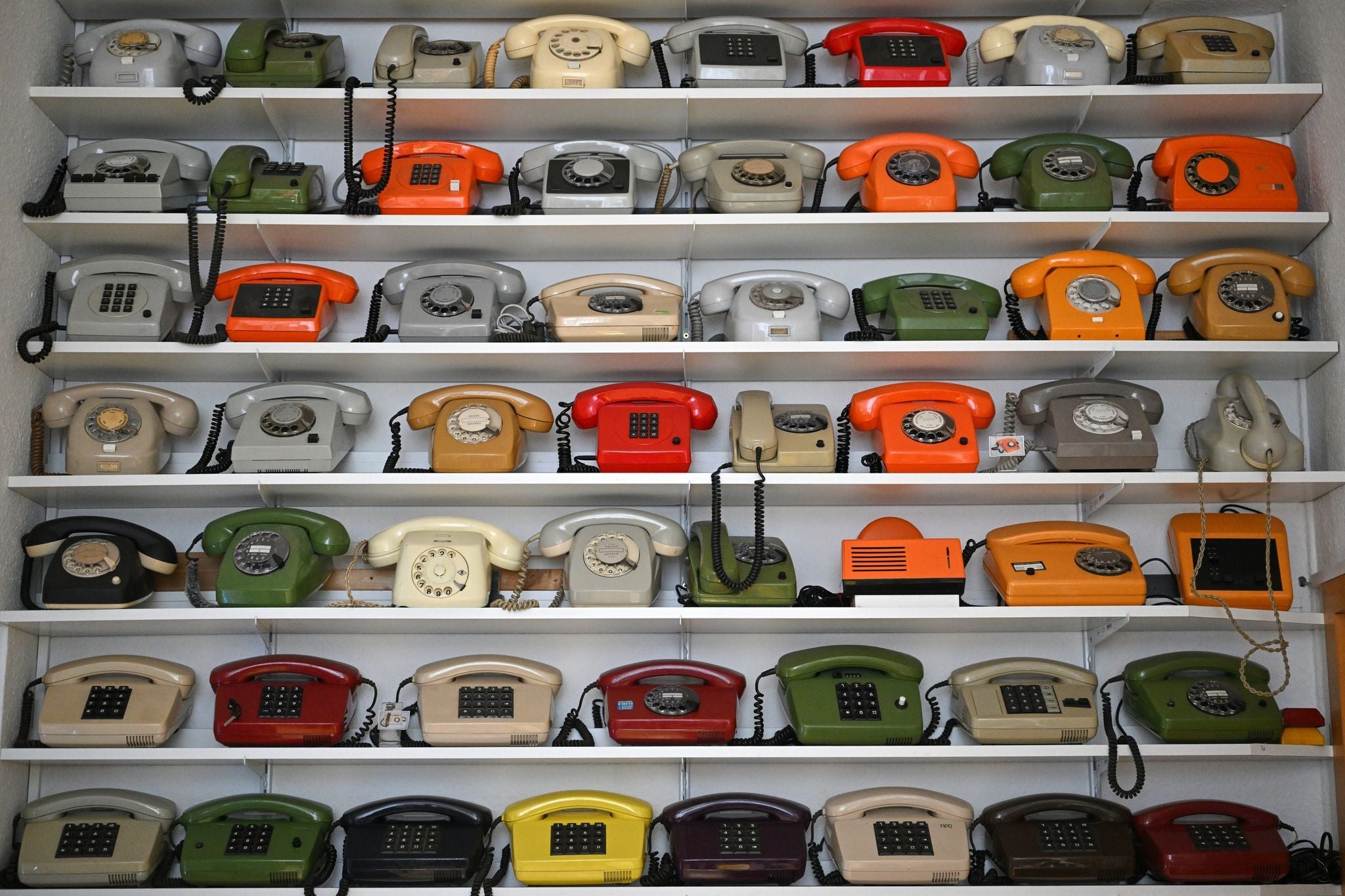 Mehrere Reihen bunter, historischer Telefone stehen auf weißen Regalbrettern im Museum.