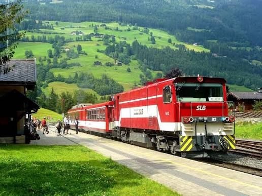 Roter Zug steht an einem kleinen Bahnhof in einer grünen, hügeligen Landschaft.