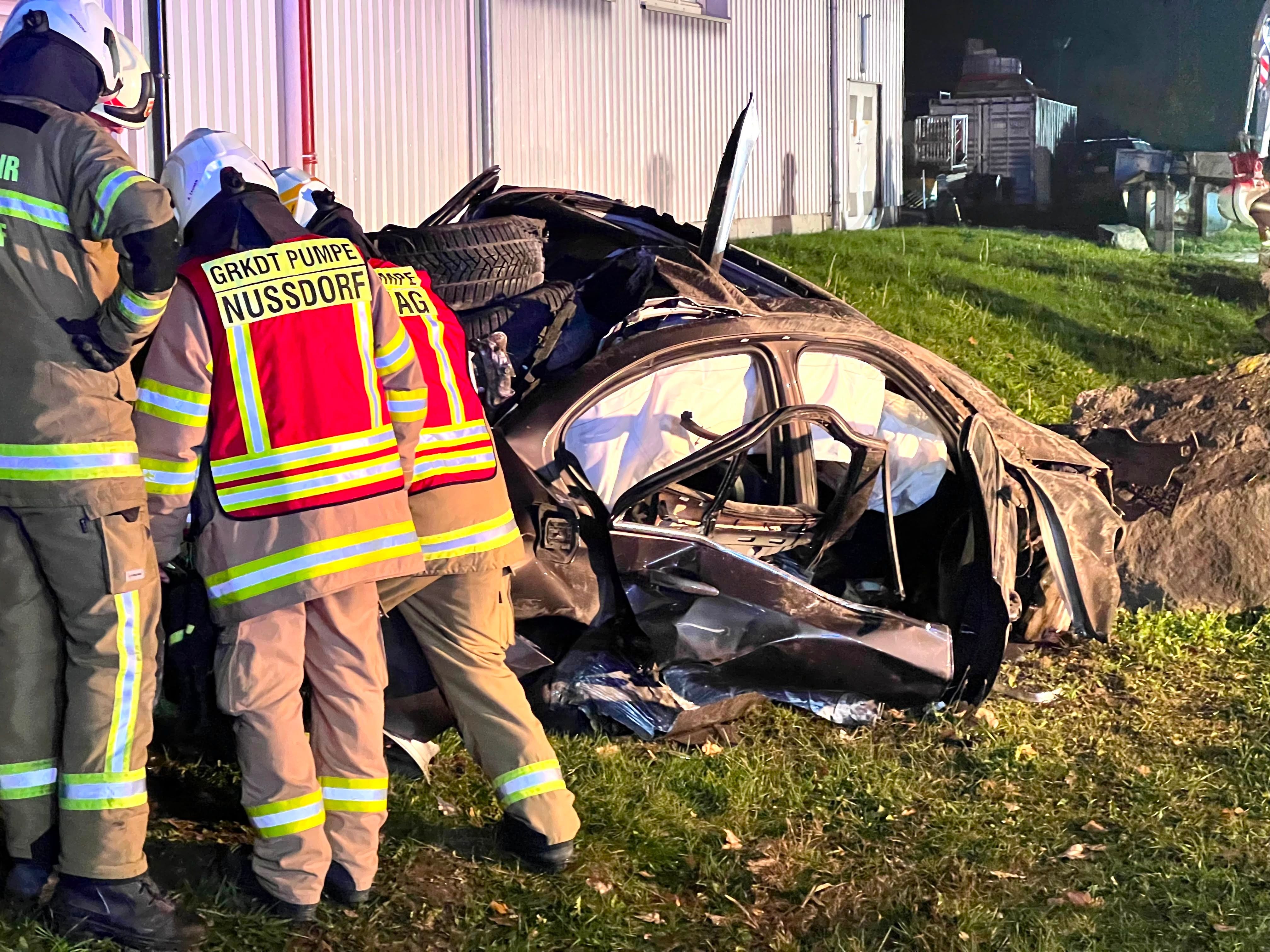 Feuerwehrleute stehen neben einem stark verunfallten und zerstörten Auto bei Nacht.