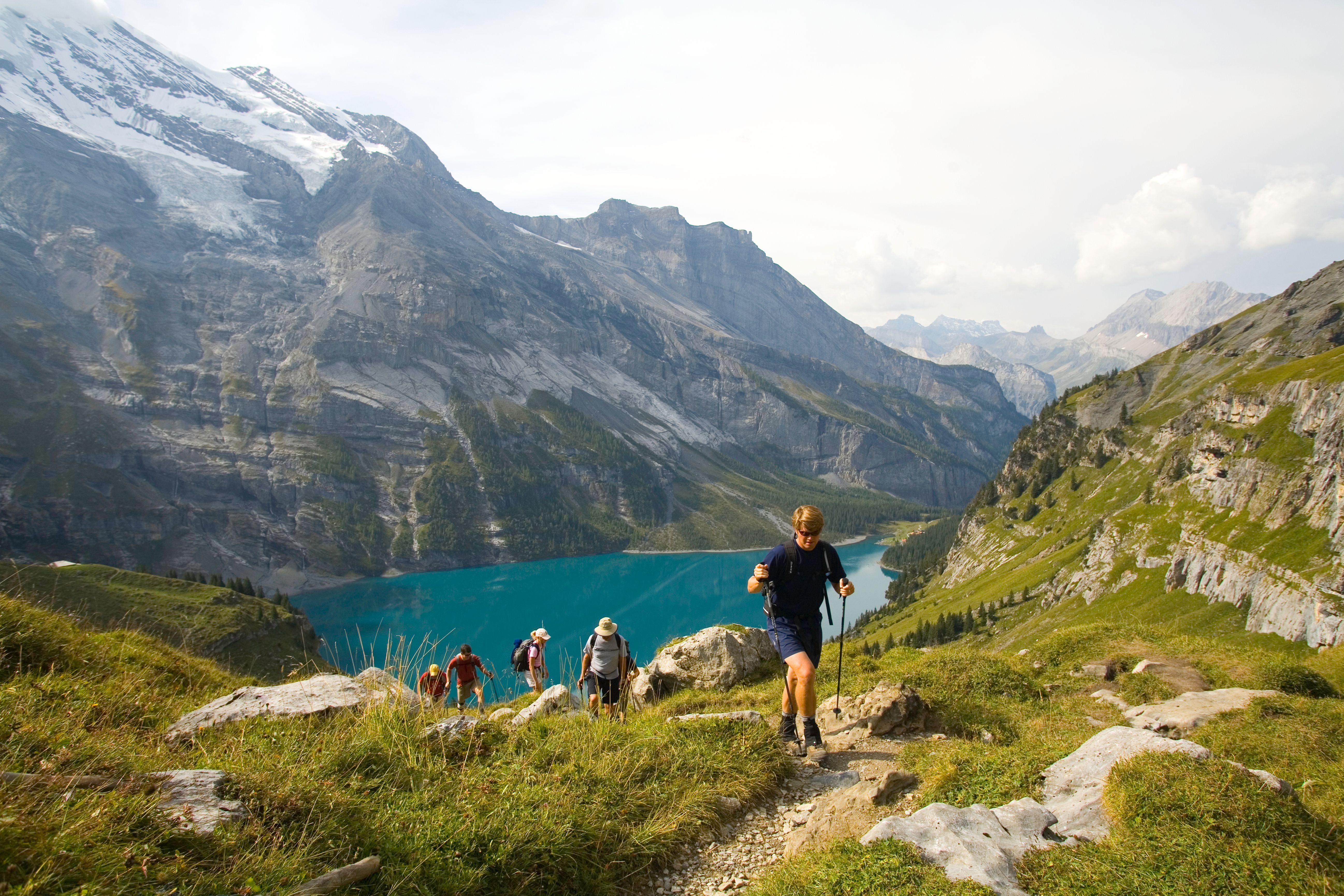 Wanderer auf einem Bergpfad mit Blick auf einen See und verschneite Berge im Hintergrund.