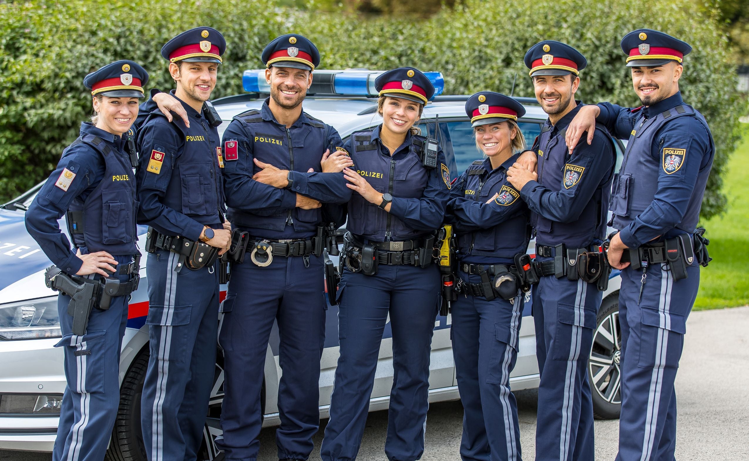 Sieben Polizisten in Uniform posieren lächelnd vor einem Polizeiauto im Freien.