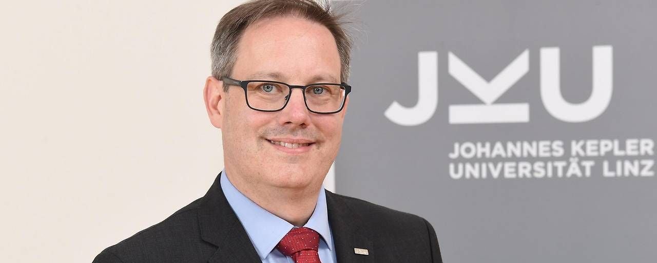 Ein Mann im Anzug steht vor einem JKU-Logo und lächelt in die Kamera.