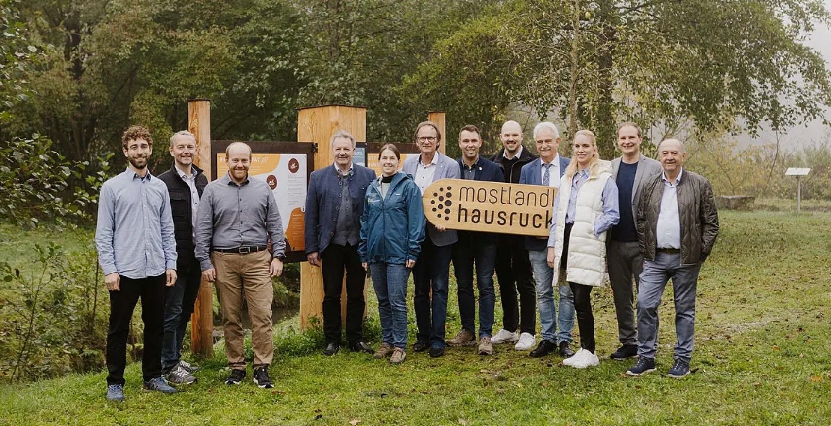 Eine Gruppe von Menschen steht im Grünen vor Infotafeln und hält ein „mostland hausruck“-Schild.