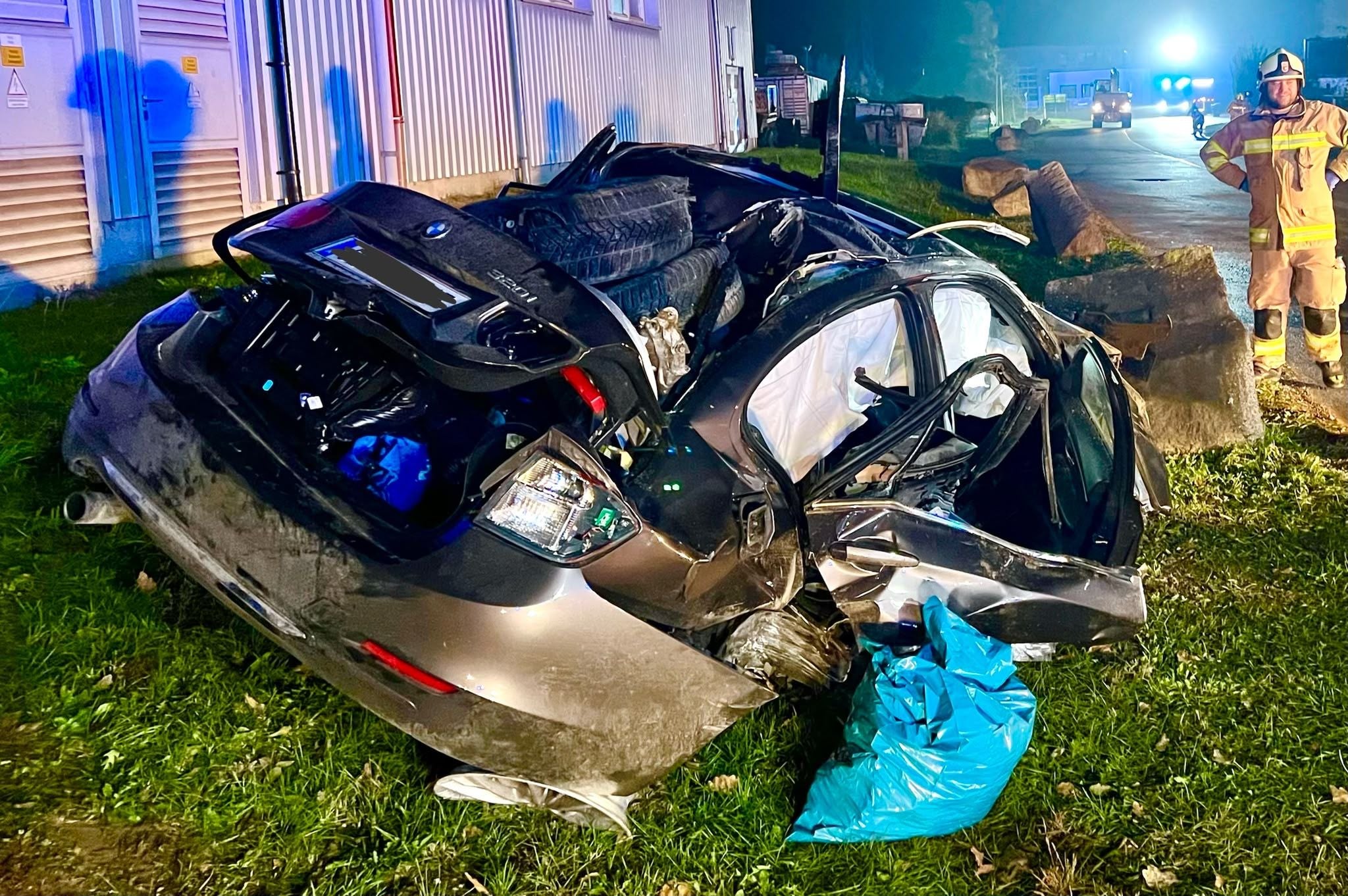 Stark zerstörtes Auto nach Unfall, Feuerwehrmann steht im Hintergrund auf einer Straße.