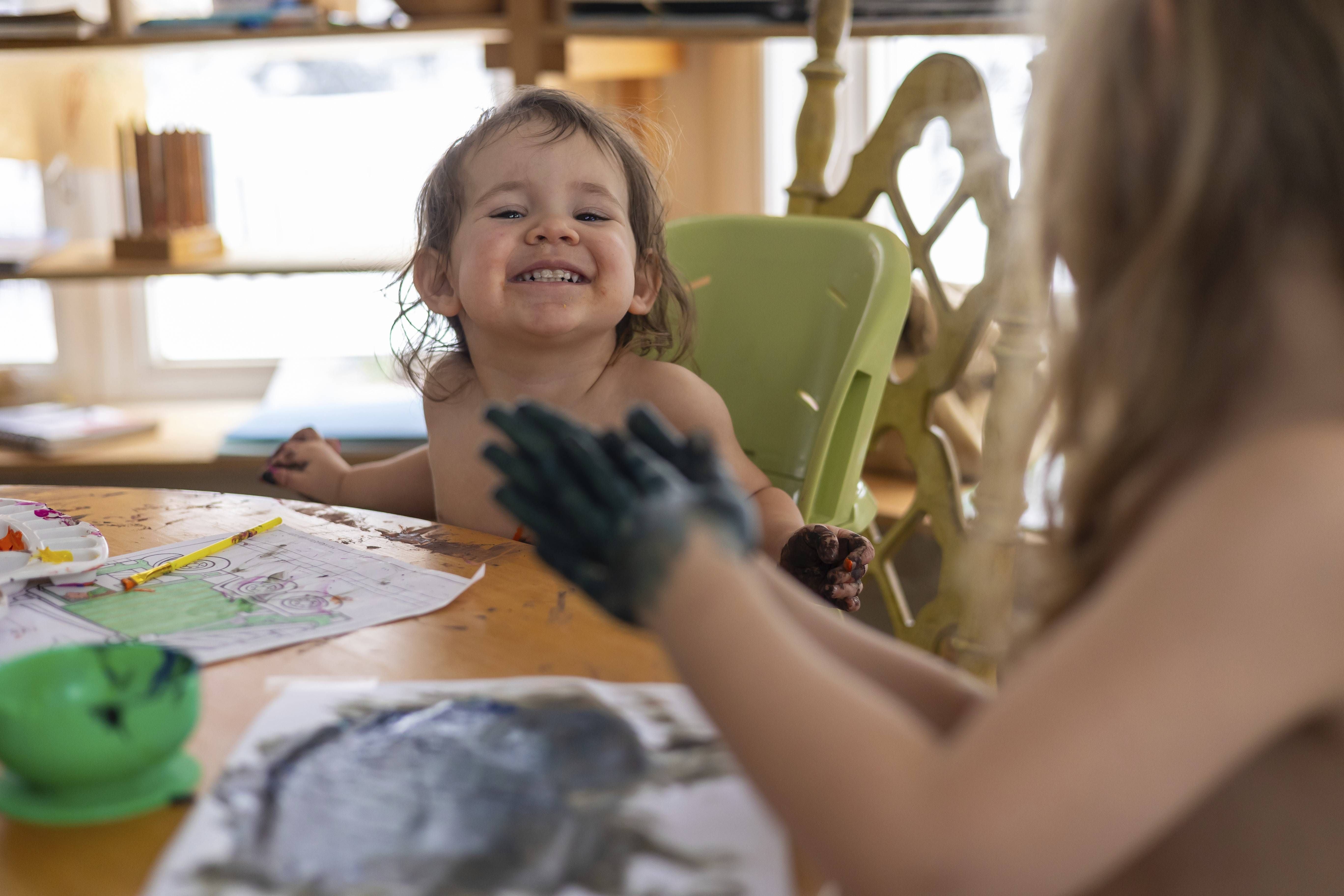 Zwei fröhliche Kinder sitzen lachend am Tisch und malen mit Farben auf Papier.