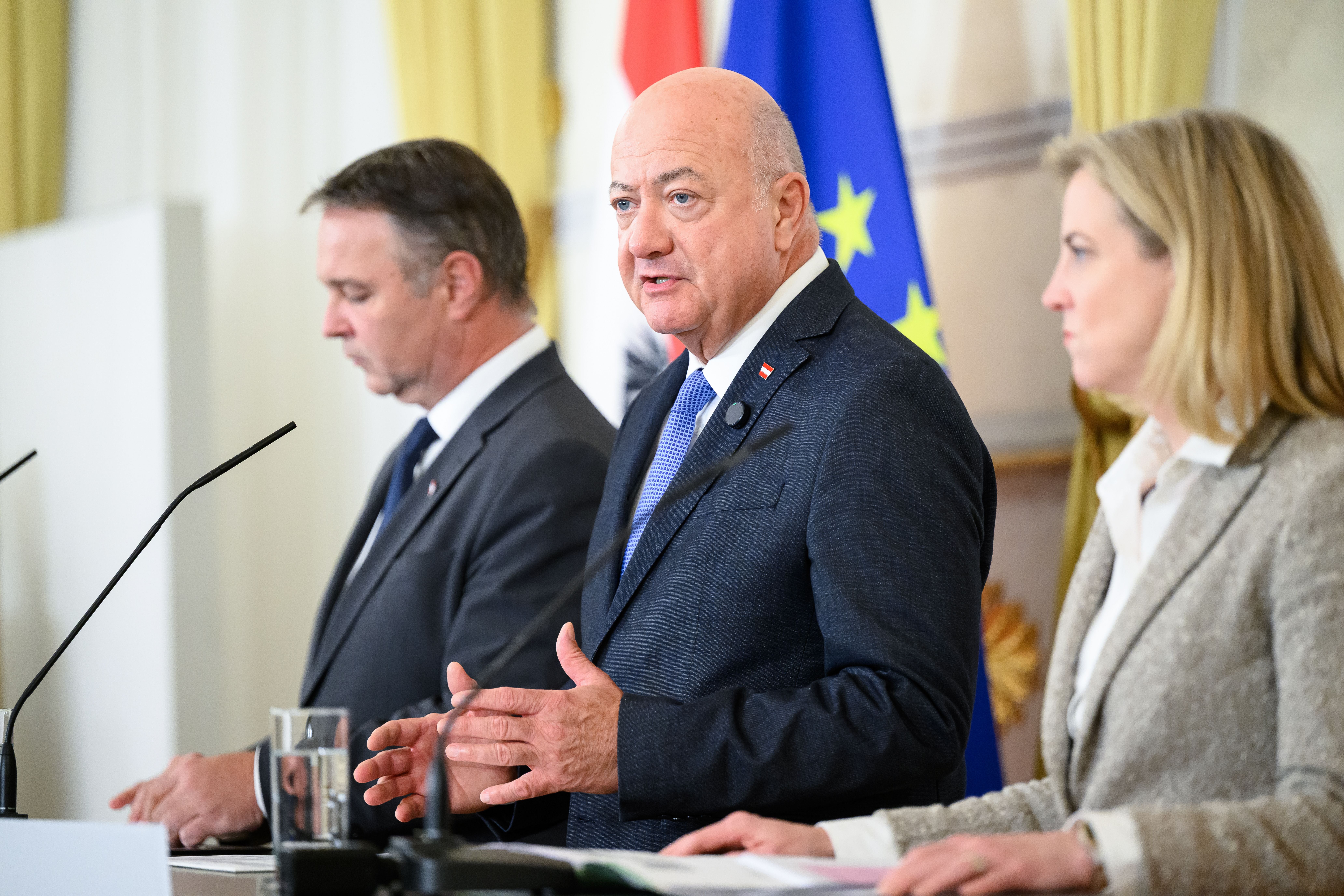 Drei Politiker sprechen bei einer Pressekonferenz vor EU- und Österreich-Flaggen in Wien.