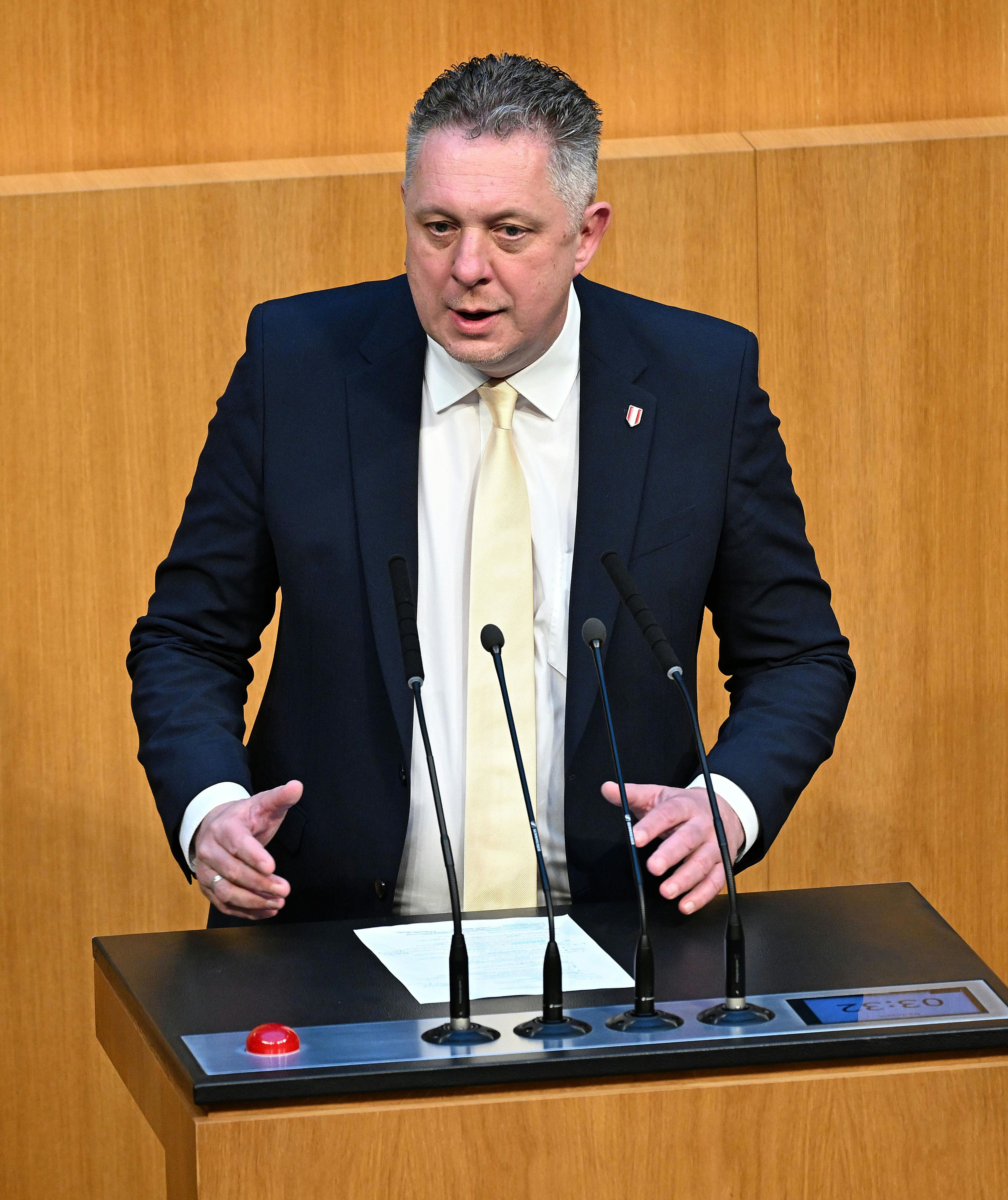 Ein Mann im Anzug spricht stehend an einem Rednerpult mit Mikrofonen im Parlament.