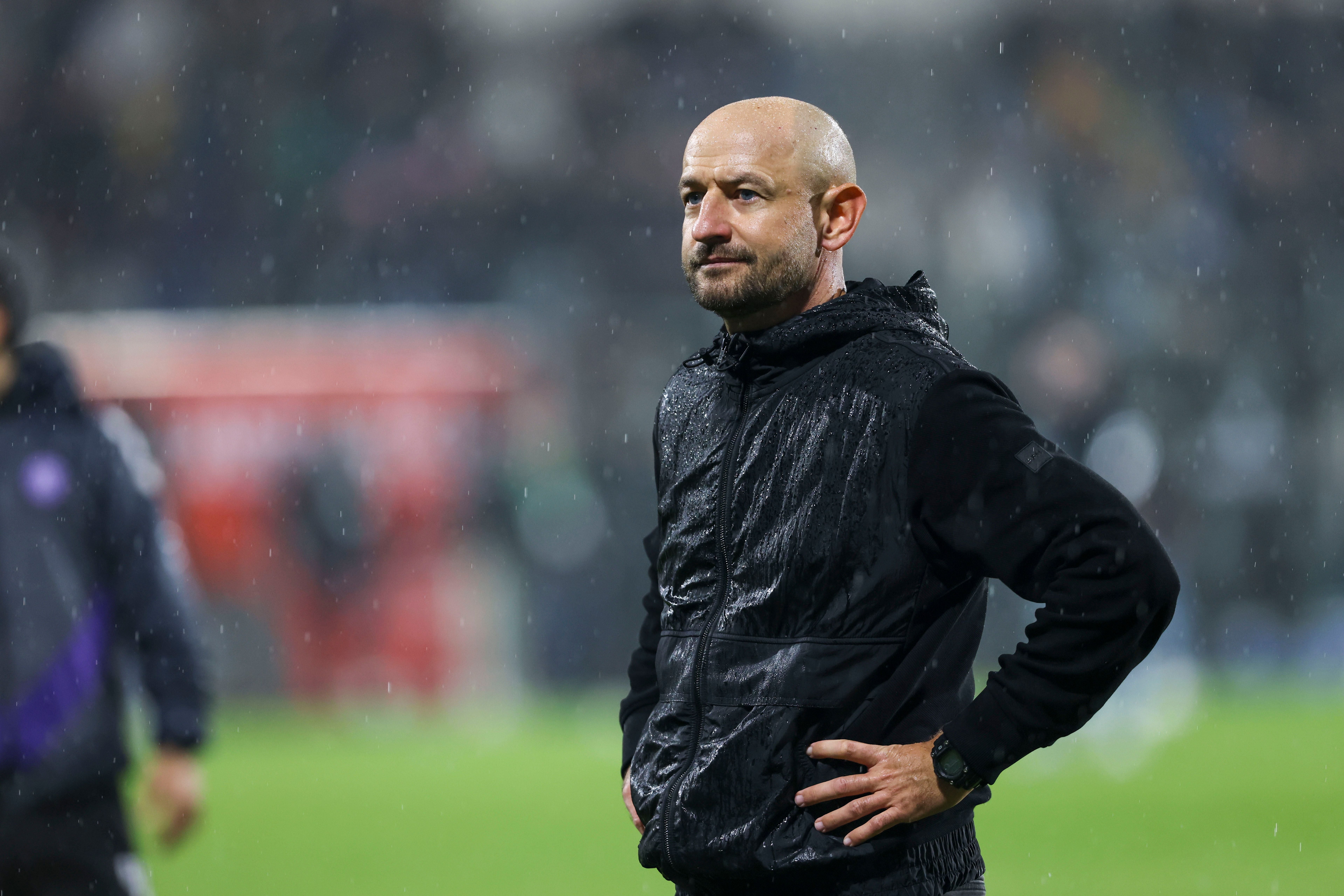 Ein Mann steht im Regen auf einem Fußballfeld und trägt eine schwarze Jacke.