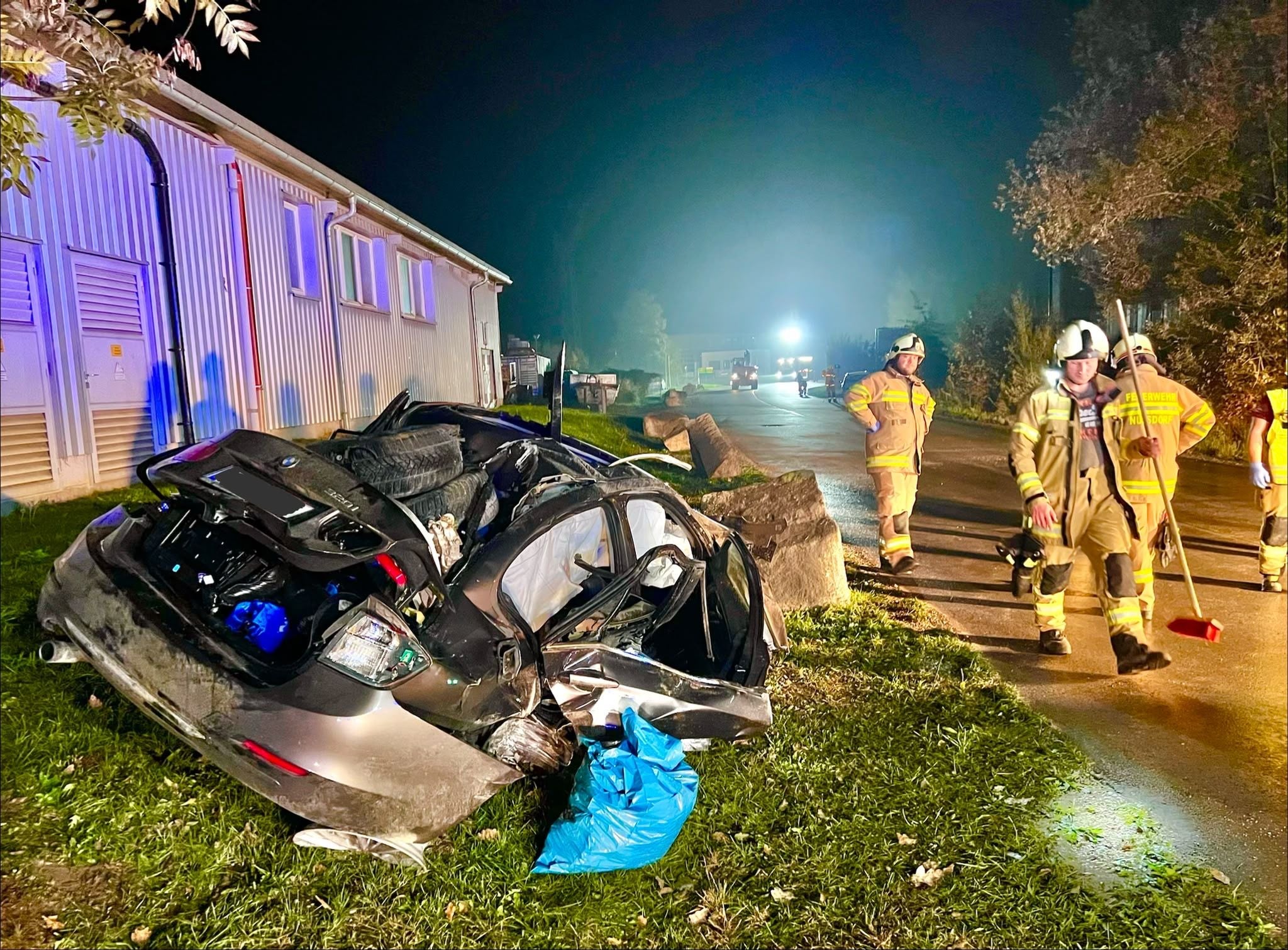 Ein schwer beschädigtes Auto nach einem Unfall, Feuerwehrleute stehen daneben auf einer nassen Straße.