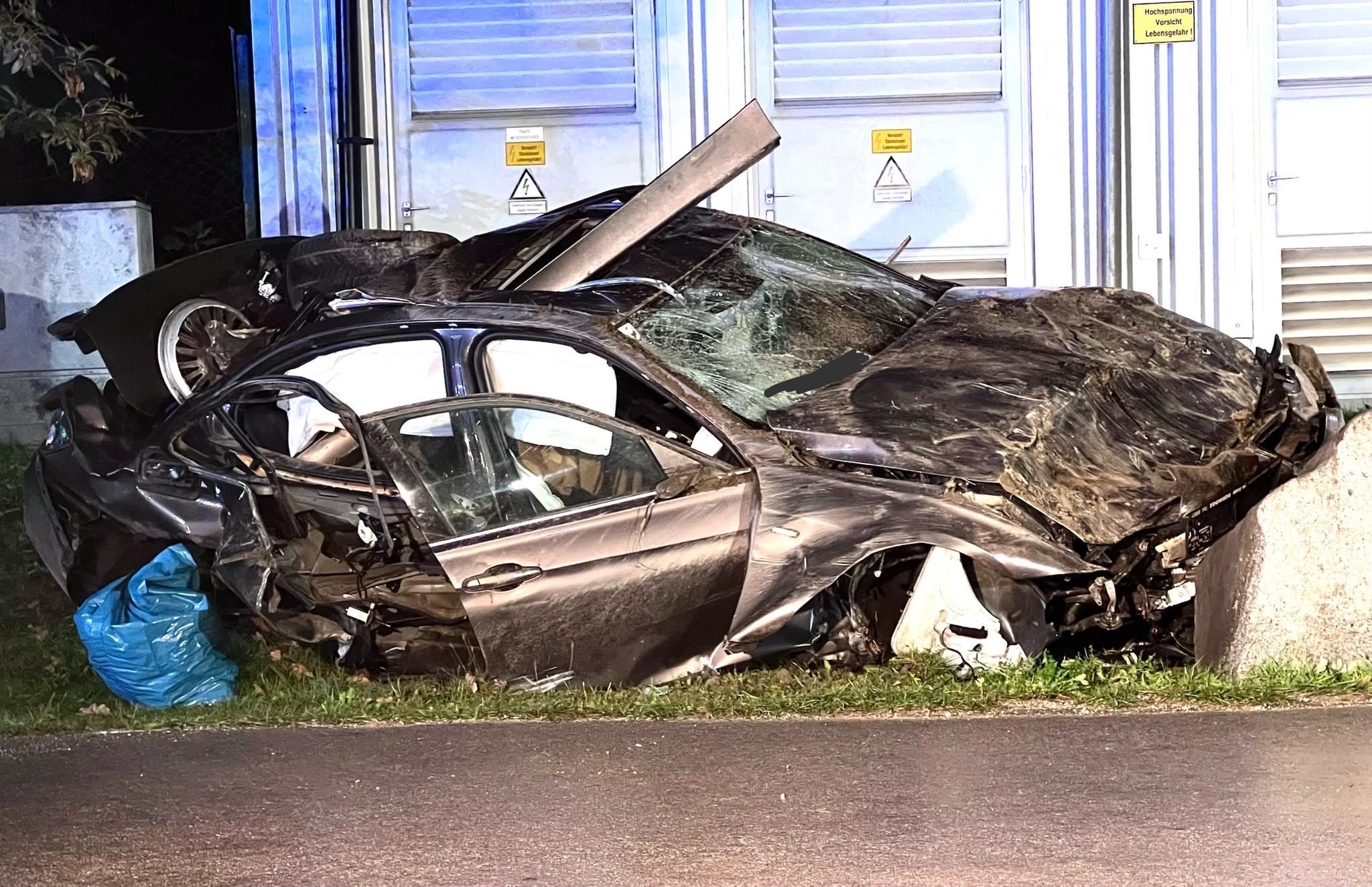 Stark beschädigtes Auto nach schwerem Unfall vor einem Gebäude mit Warnschildern.