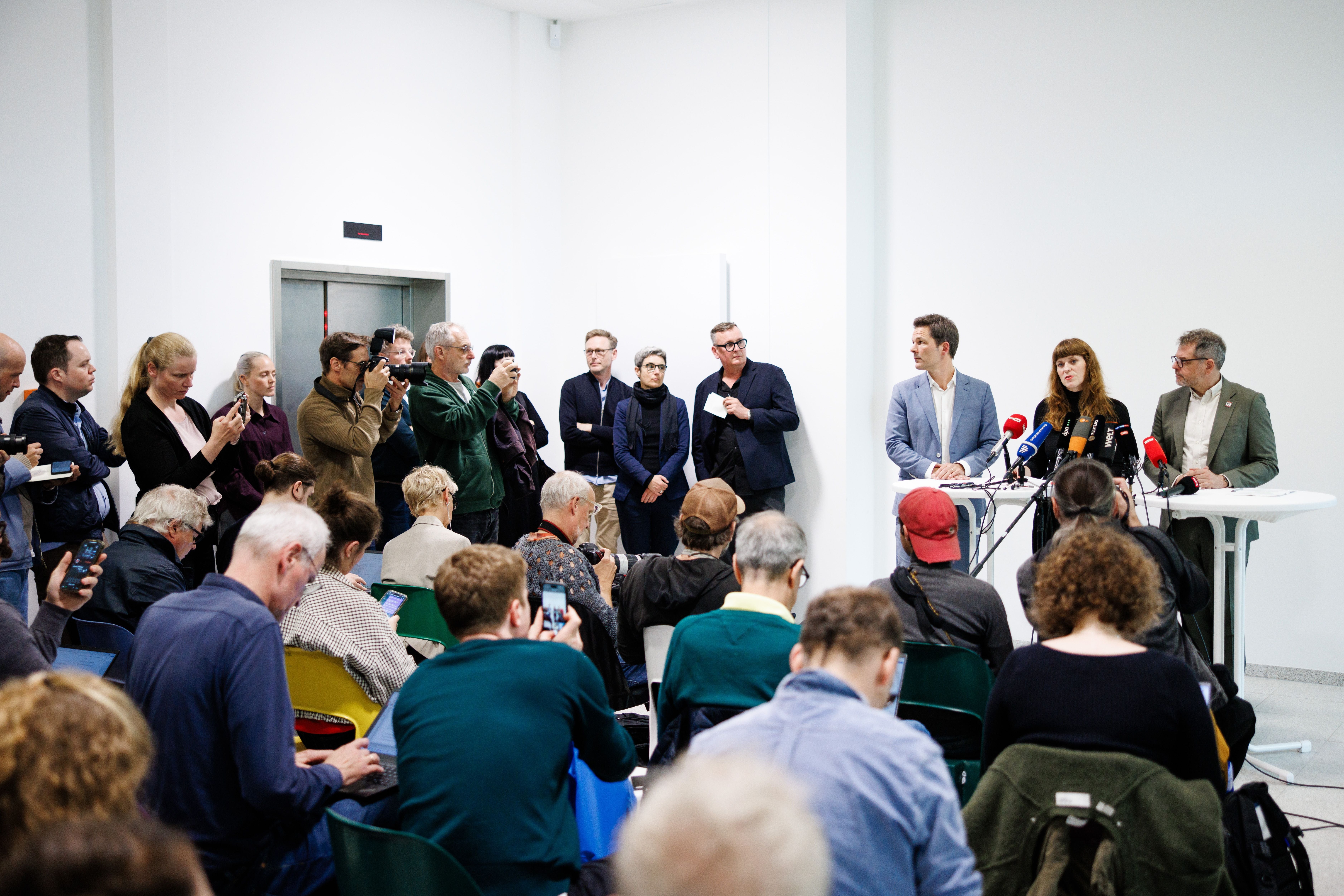 Pressekonferenz mit Politikern, viele Journalisten fotografieren und machen Notizen im Raum.