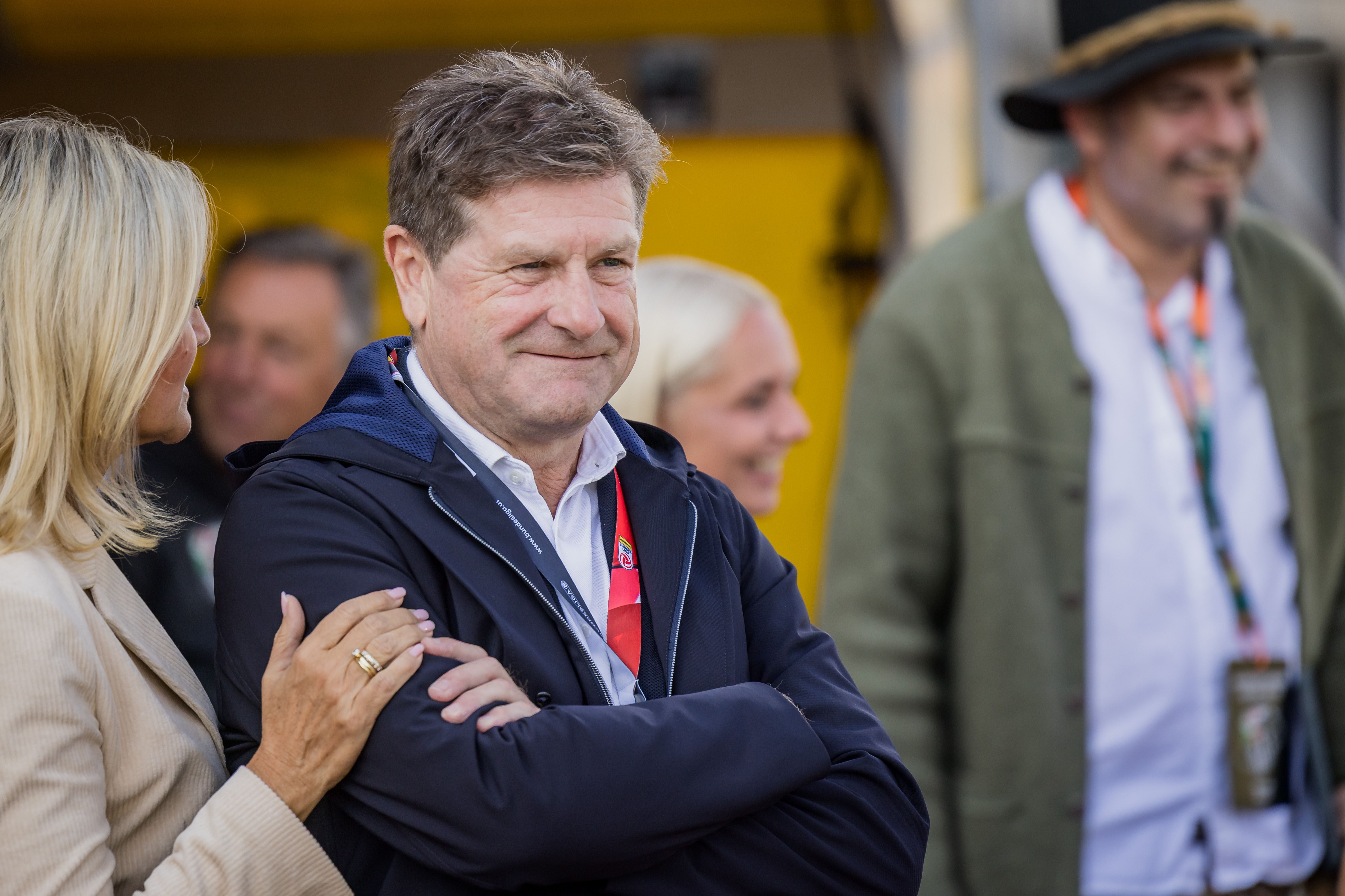 Ein Mann steht mit verschränkten Armen und lächelt, weitere Personen im Hintergrund.