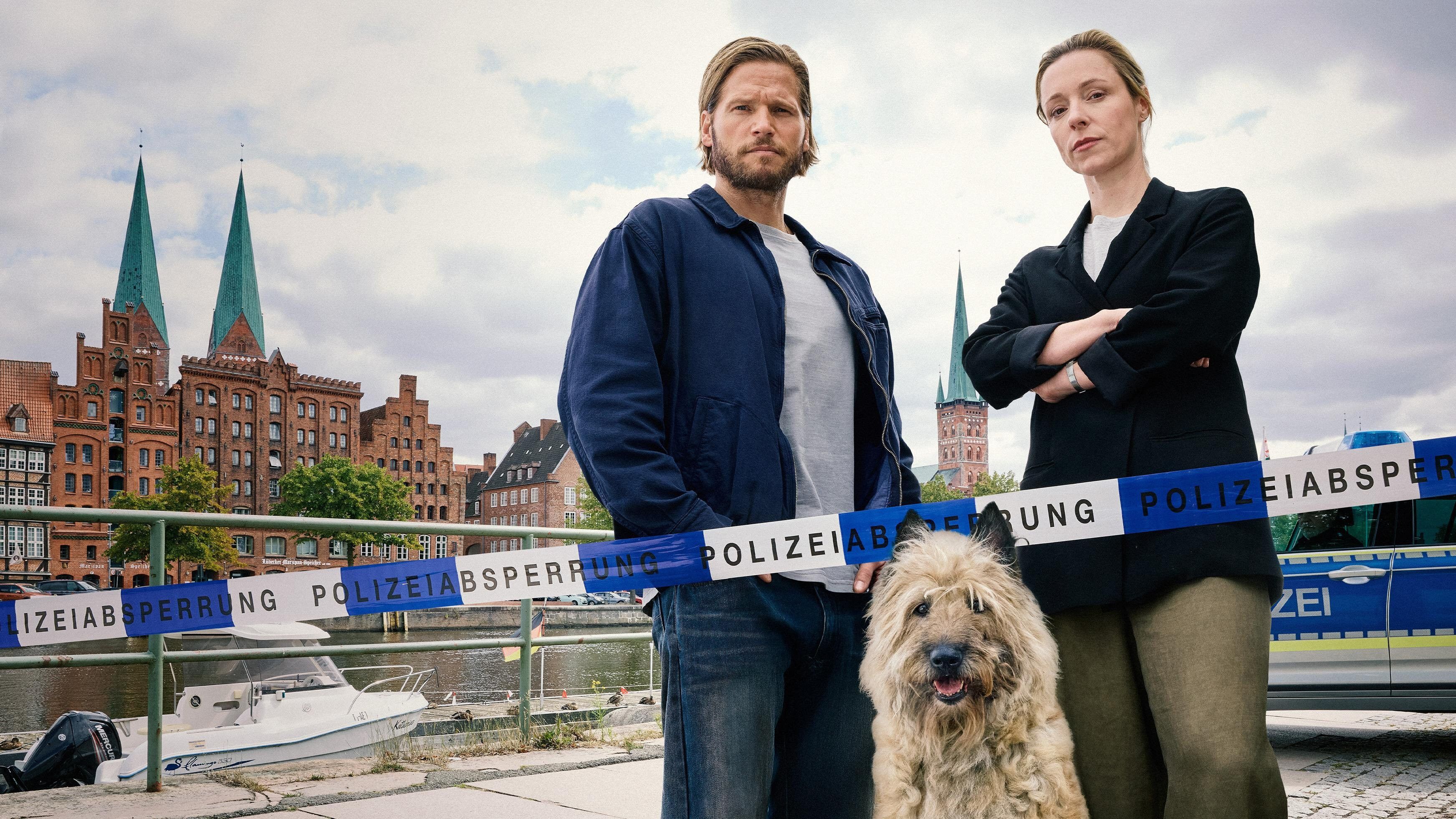 Zwei Personen und ein Hund stehen an einem Polizeiband vor historischer Lübecker Kulisse am Wasser.