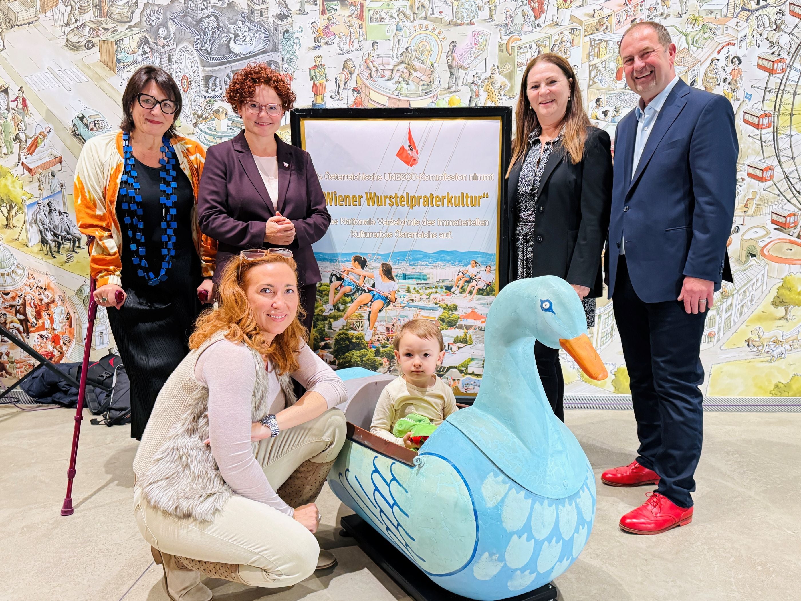 Fünf Erwachsene posieren mit einem Kind in einer blauen Entenfigur vor einem Plakat zur Praterkultur.