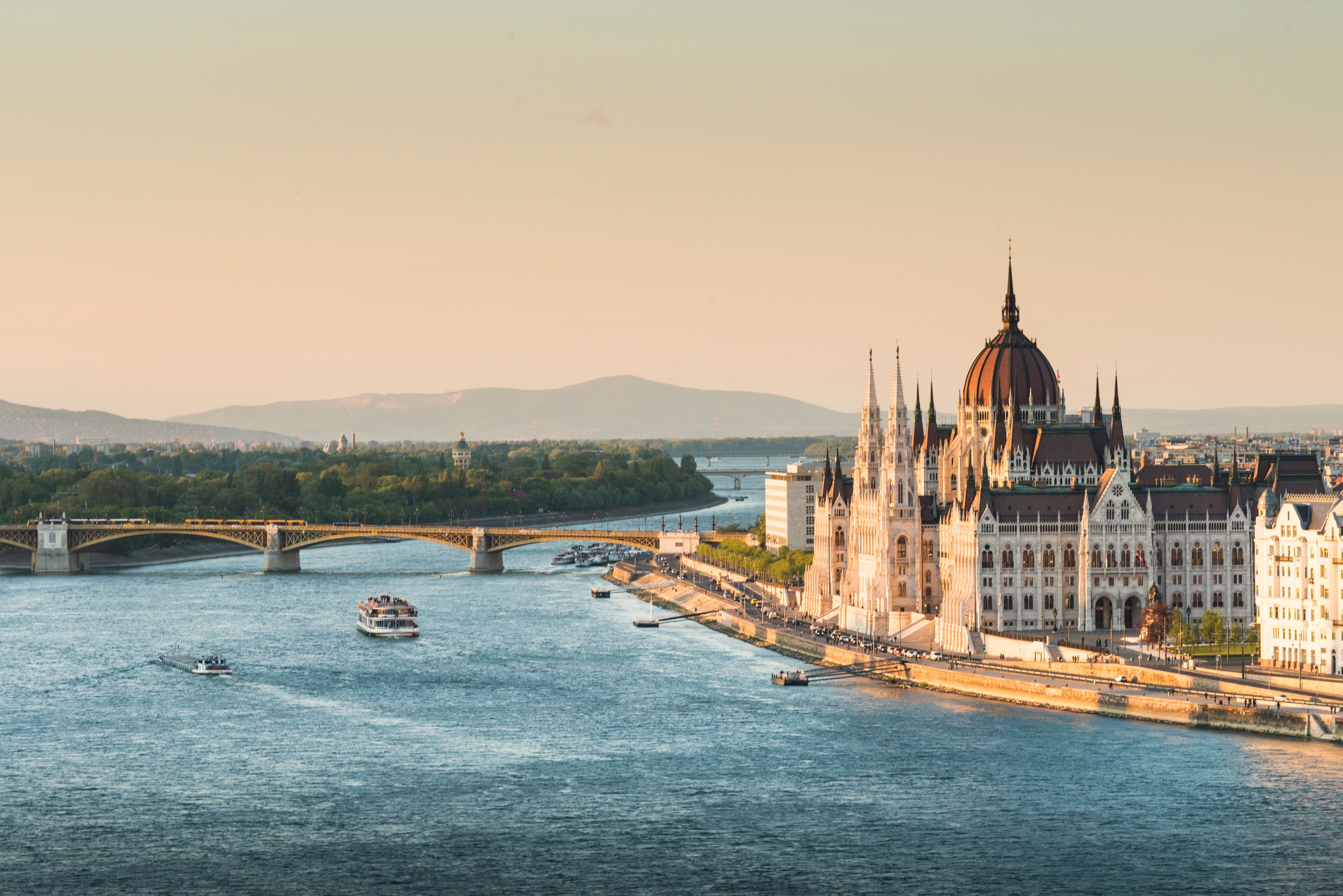 Das ungarische Parlament am Donauufer in Budapest bei Sonnenuntergang, mit Schiffen auf dem Fluss.
