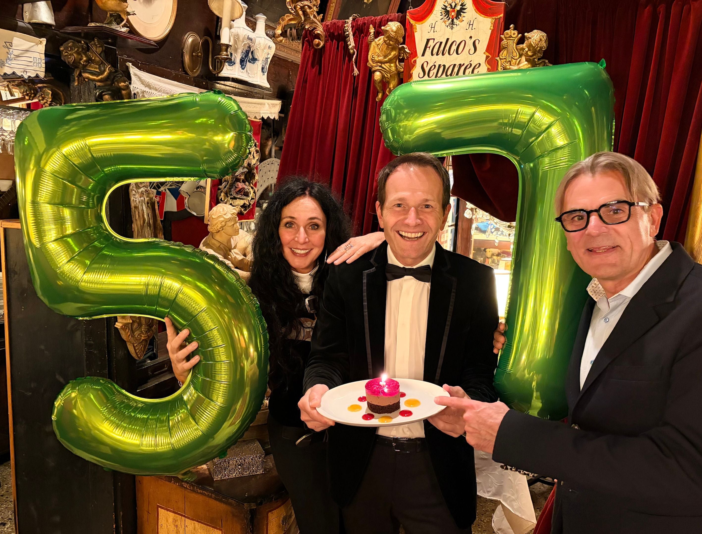 Drei Personen feiern mit grünen Luftballons in Form der Zahl 57 und einem kleinen Geburtstagskuchen.