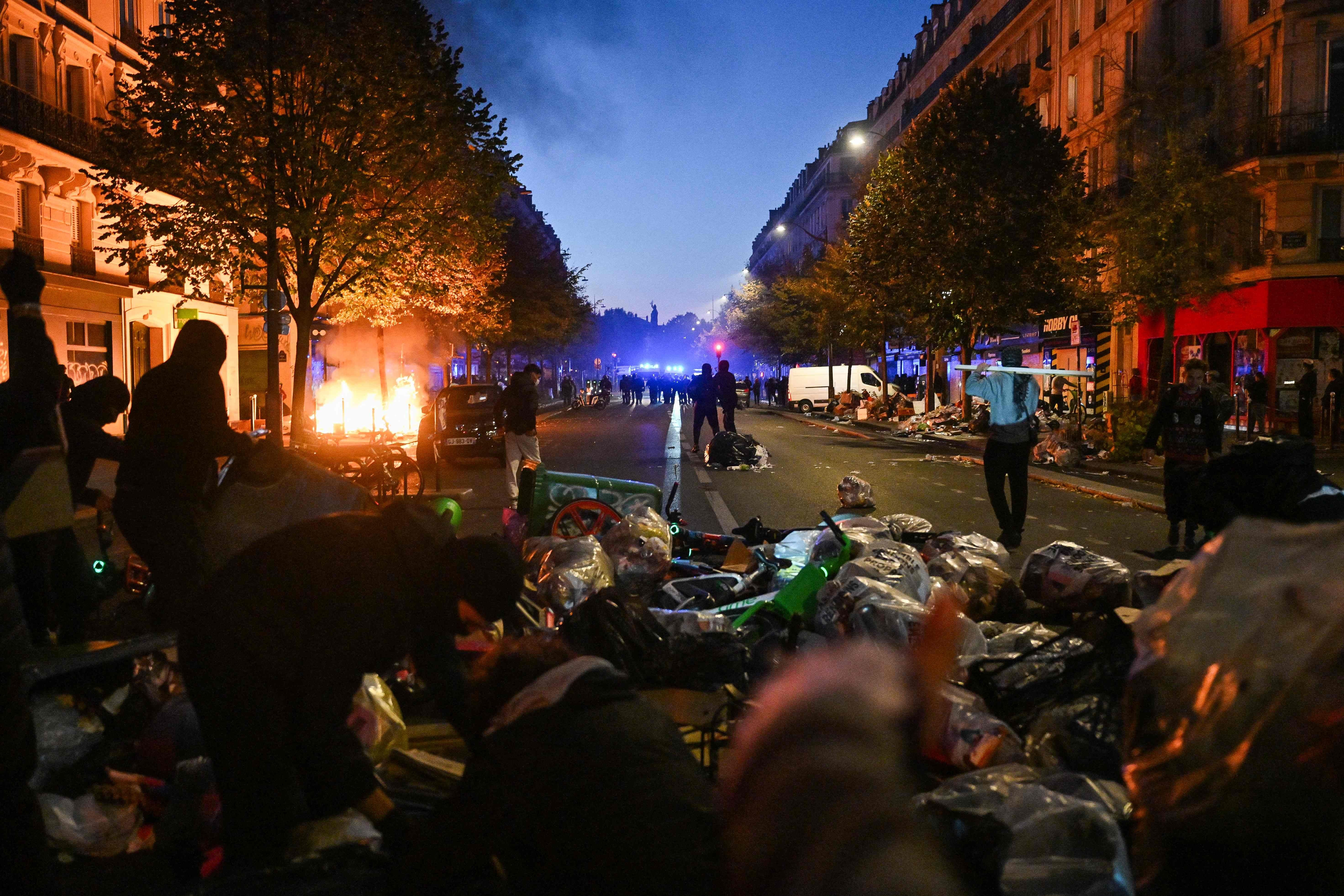 Menschen verbarrikadieren eine Straße in Paris mit Müll und Fahrrädern, im Hintergrund brennt etwas.