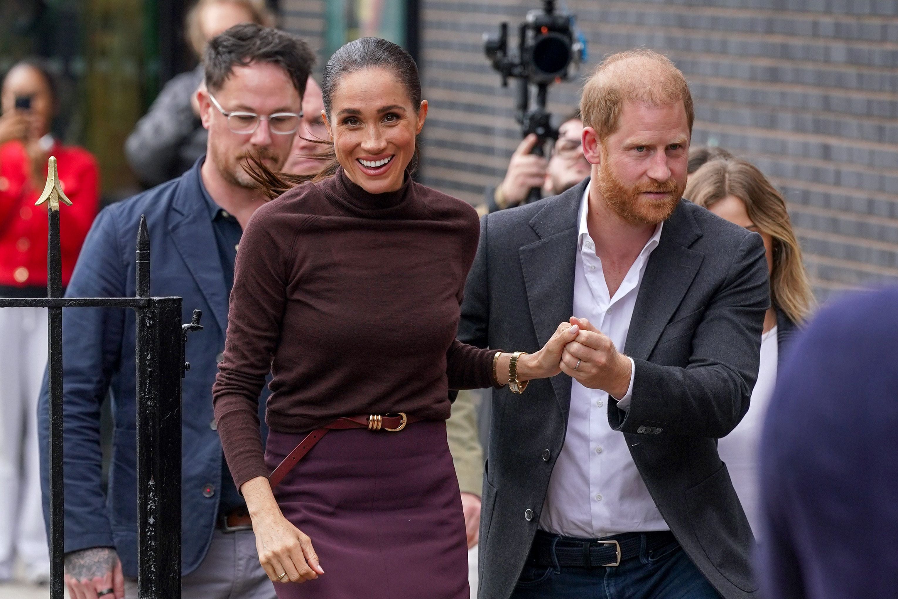 Meghan Markle und Prinz Harry gehen lächelnd Hand in Hand durch ein Tor, umgeben von weiteren Personen.