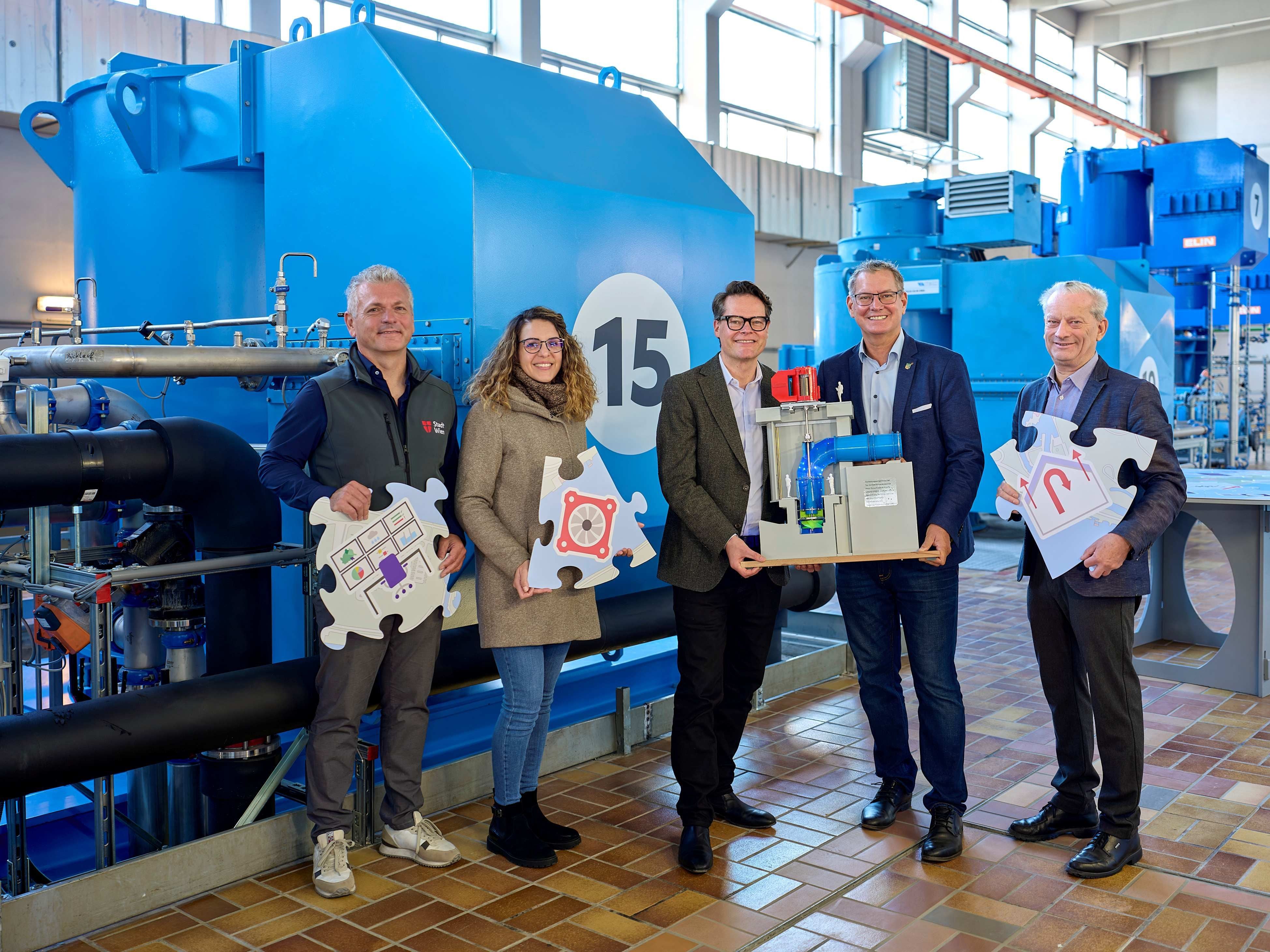 Fünf Personen stehen vor blauen Maschinen und halten Puzzleteile zum Thema Überflutungsschutz.